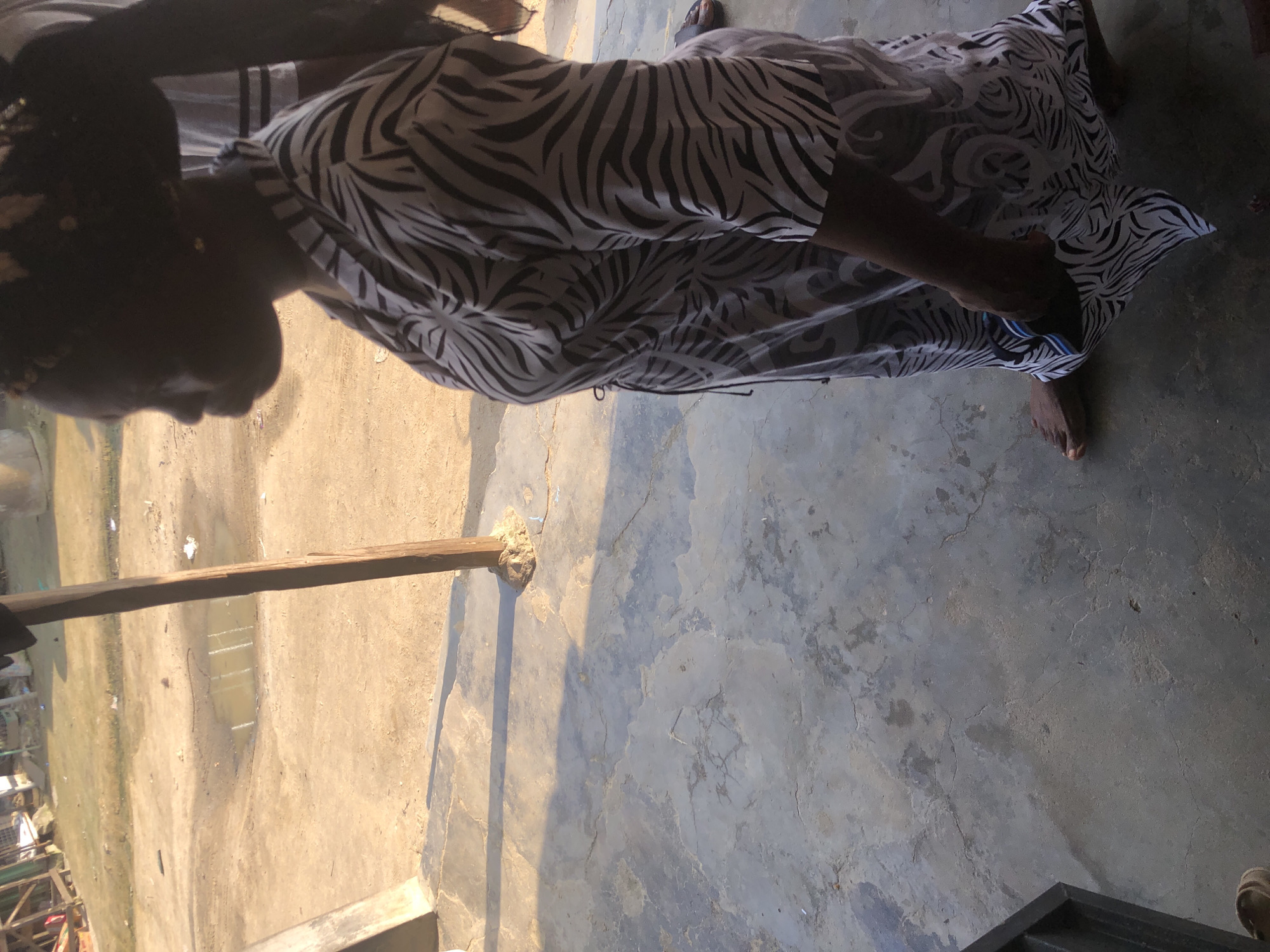 A side profile image shows a person from the chest down, standing on a cracked concrete surface, partially shaded by what appears to be an overhang. The person is wearing a long, flowing dress with a black and white zebra-like pattern. The dress has a cinched waist and wide sleeves. A dark cord hangs from the waistline. The person's bare feet are visible at the bottom of the frame, with one foot slightly forward. In the background, a dirt or sandy area is visible, with a shallow puddle of water. Further back, wooden structures and sparse vegetation can be seen. The lighting suggests it is daytime, with harsh shadows cast on the concrete.