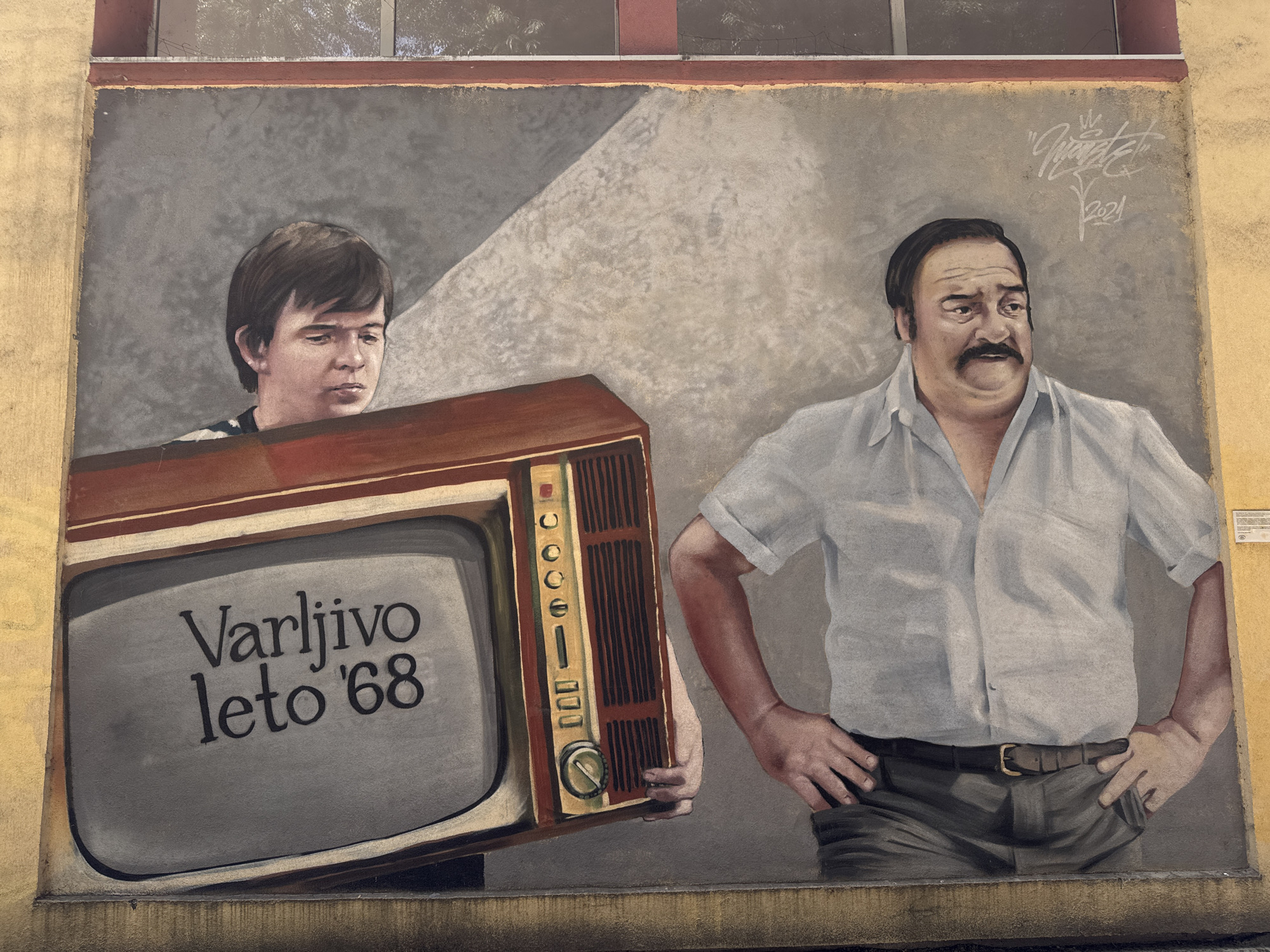 The image shows a mural on a wall in Novi Sad, Serbia. The mural depicts a young man holding an old-fashioned television set displaying the text "Varljivo leto '68," a popular Yugoslav film title. Standing next to the young man is an older man with a mustache, wearing a short-sleeved shirt and pants, with his hands on his hips. The background is a mottled gray with a beam of light. The painting is signed with the artist's signature and the date "2021" in the top right corner. The edges of the mural blend into the wall upon which it is painted. There is a partially obscured window above the mural. The overall style suggests a realistic depiction.