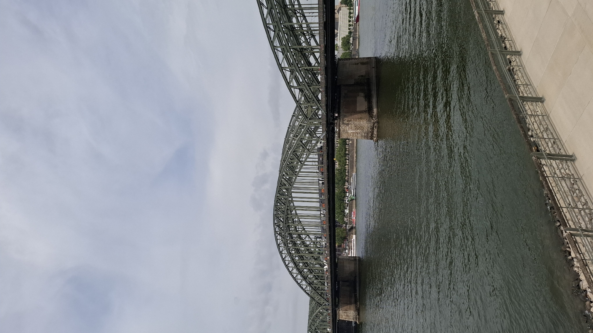 An outdoor, medium-shot, vertical photograph shows a view of the Hohenzollern Bridge in Cologne, Germany. The bridge is made of dark green metal with large arching structures and is supported by stone piers. Trains are visible on the bridge's deck. Below the bridge, a wide, dark green river flows. Two boats, one white and one with red accents, are on the river. The riverbank in the foreground has a paved walkway with a metal railing and is lined with stones. The background shows a treelined embankment and buildings. The sky is overcast with light gray clouds.