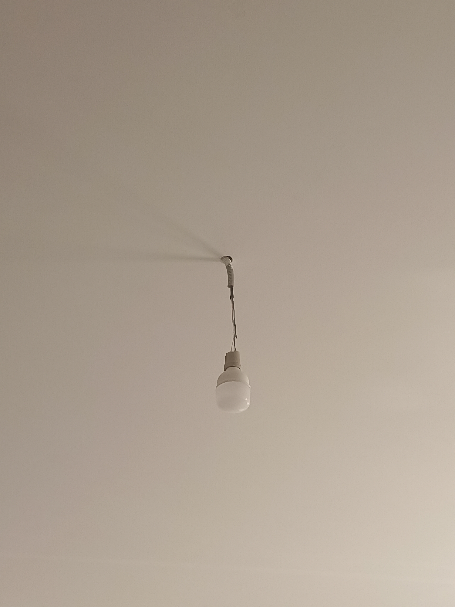 The image shows a simple ceiling with a single light bulb hanging from a wire. The light bulb is white and appears to be an LED bulb. The ceiling is a neutral color, possibly white or off-white. There is a shadow cast on the ceiling by the light bulb and its fixture. The image is taken from a low angle, looking up at the ceiling. The location is Izberbash, Russia.