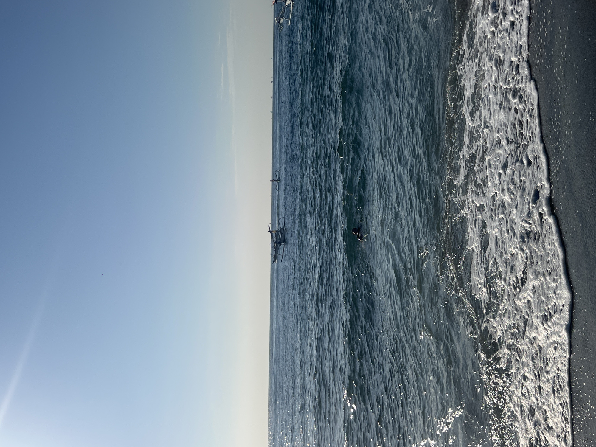 In Abang, Indonesia, on a bright day, the scene is set by the deep blue ocean and a clear sky. Waves gently lap at the dark, wet sand in the foreground, their white foam creating a stark contrast. Further out, the water glitters under the sun. Several outrigger boats are visible on the horizon, some carrying people, while a lone swimmer is present in the water closer to the shore. The environment is characteristic of a coastal area with calm seas and sunny weather.