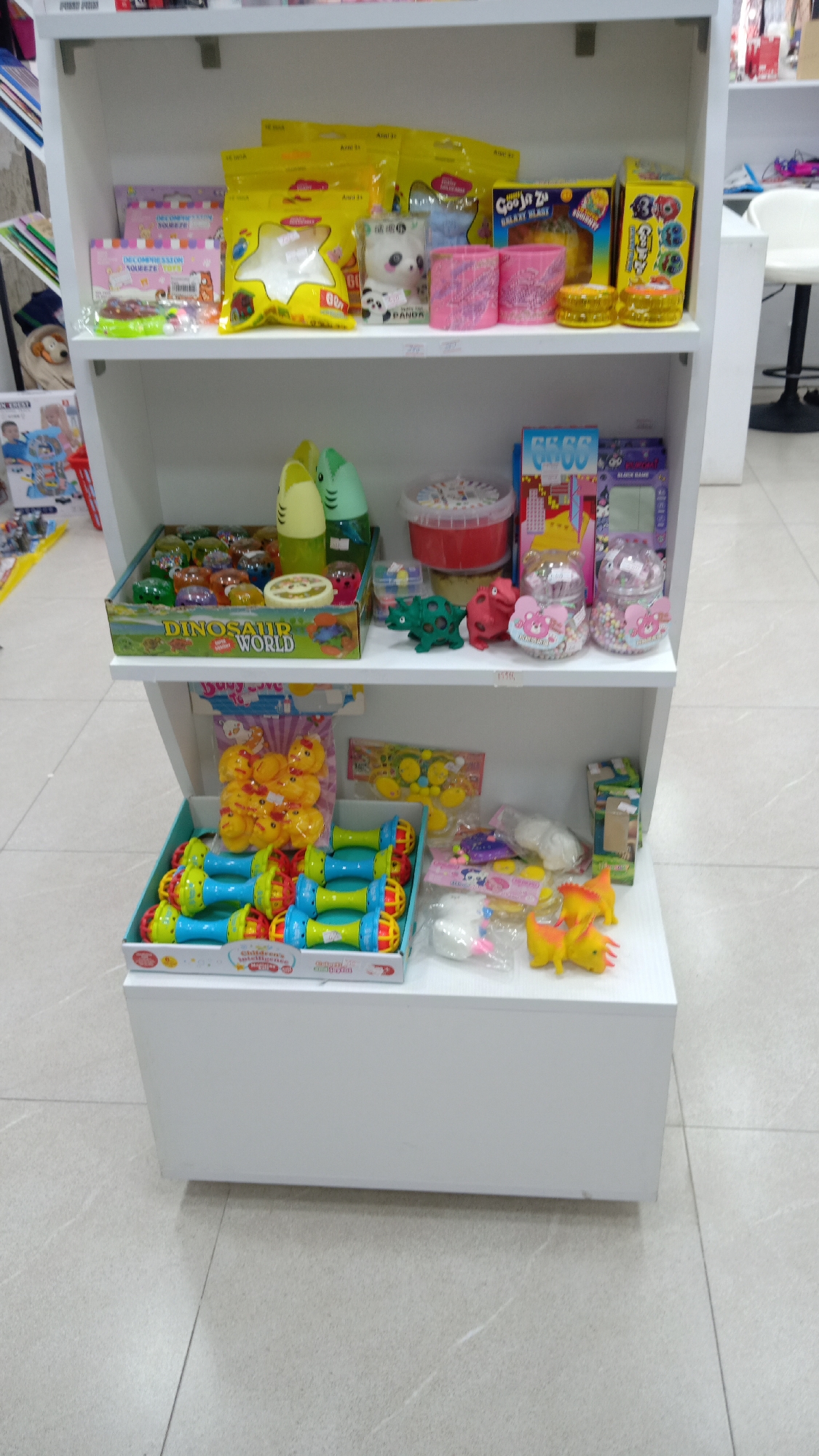 A vertically-oriented eye-level shot depicts a three-tiered white shelf unit stocked with various toys, situated on a tile floor.
On the top shelf are squeeze toys, including yellow star-shaped toys in clear packaging. A white and black panda plush toy, pink playdough, and Goo Jit Zu Galaxy Blast toys are also present.
The middle shelf features a "Dinosaur World" box filled with dinosaur toys, a container with a red lid, and card games. Clear containers filled with small candies are also visible.
The bottom shelf showcases yellow rubber ducks, blue and green dumbbell-shaped toys, and more toys wrapped in clear plastic.
The shelves are placed on a white storage unit on the floor. The tile flooring is uniform throughout the scene.