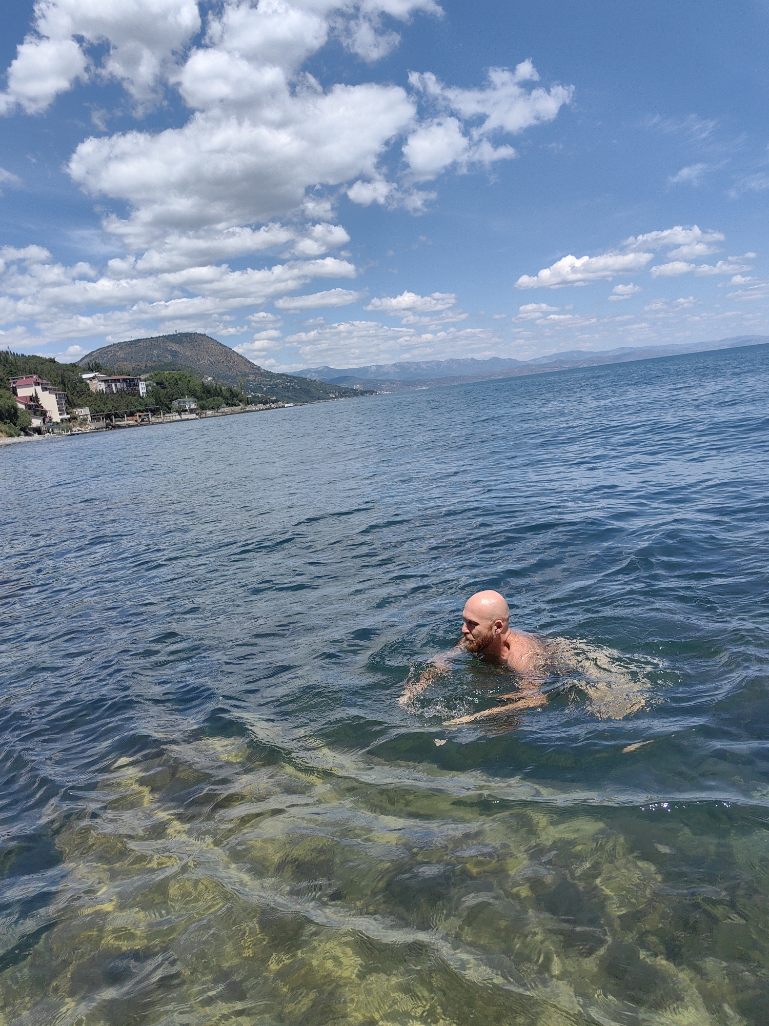 A bald, middle-aged Caucasian man with a beard is swimming in the calm, blue waters of a large body of water, possibly a lake or sea. The water is clear enough in the foreground to see the rocky, yellowish-green bottom. The man's head and shoulders are visible as he swims, his arms partially submerged. In the background, a coastline is visible with buildings and lush green trees on a hillside, leading up to a prominent, conical hill. A hazy mountain range can be seen in the far distance. The sky is a vibrant blue, dotted with fluffy white clouds. The location is identified as Utios, Ukraine.