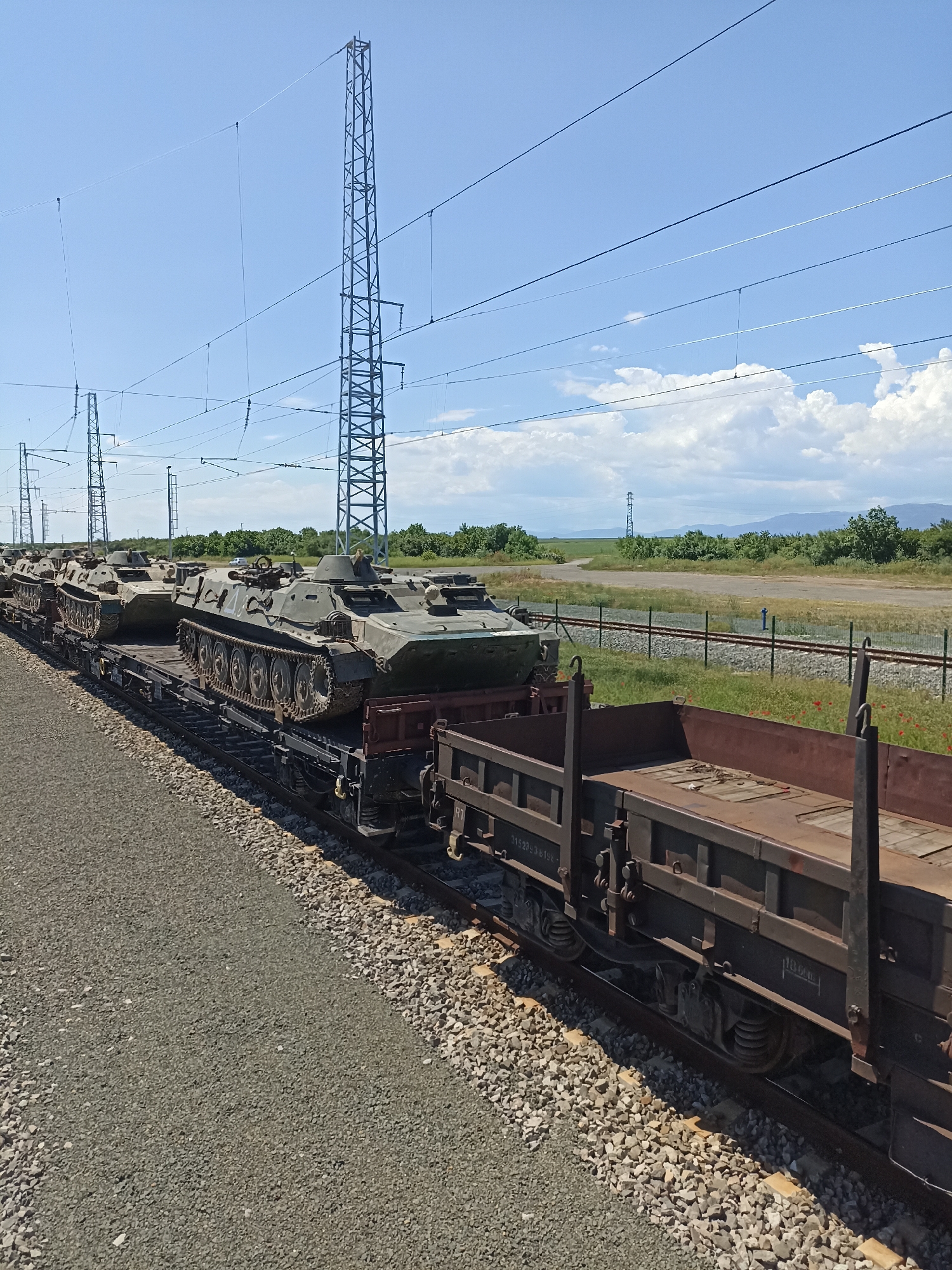 The image depicts a train carrying several armored fighting vehicles, likely BMPs, traveling on a railway line. The train is composed of flatbed cars and gondola cars. The surrounding environment features an open landscape with gravel ground, power lines supported by tall metal pylons, a fence, and some vegetation. In the background, there are hills and a partly cloudy sky. While not directly discernible from the image, the location is purportedly Zimnitsa, Bulgaria.