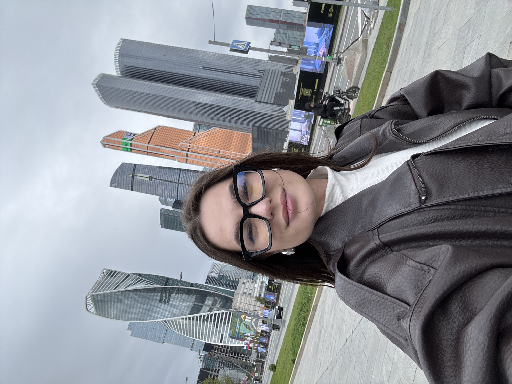 A young woman poses for a selfie in Moscow, Russia. She is wearing a brown leather jacket and glasses, standing on a sidewalk with tall, modern skyscrapers in the background. The "Evolution Tower" is on the left. A crosswalk sign is in the background, with a motorcyclist parked behind it. The sky is overcast.
