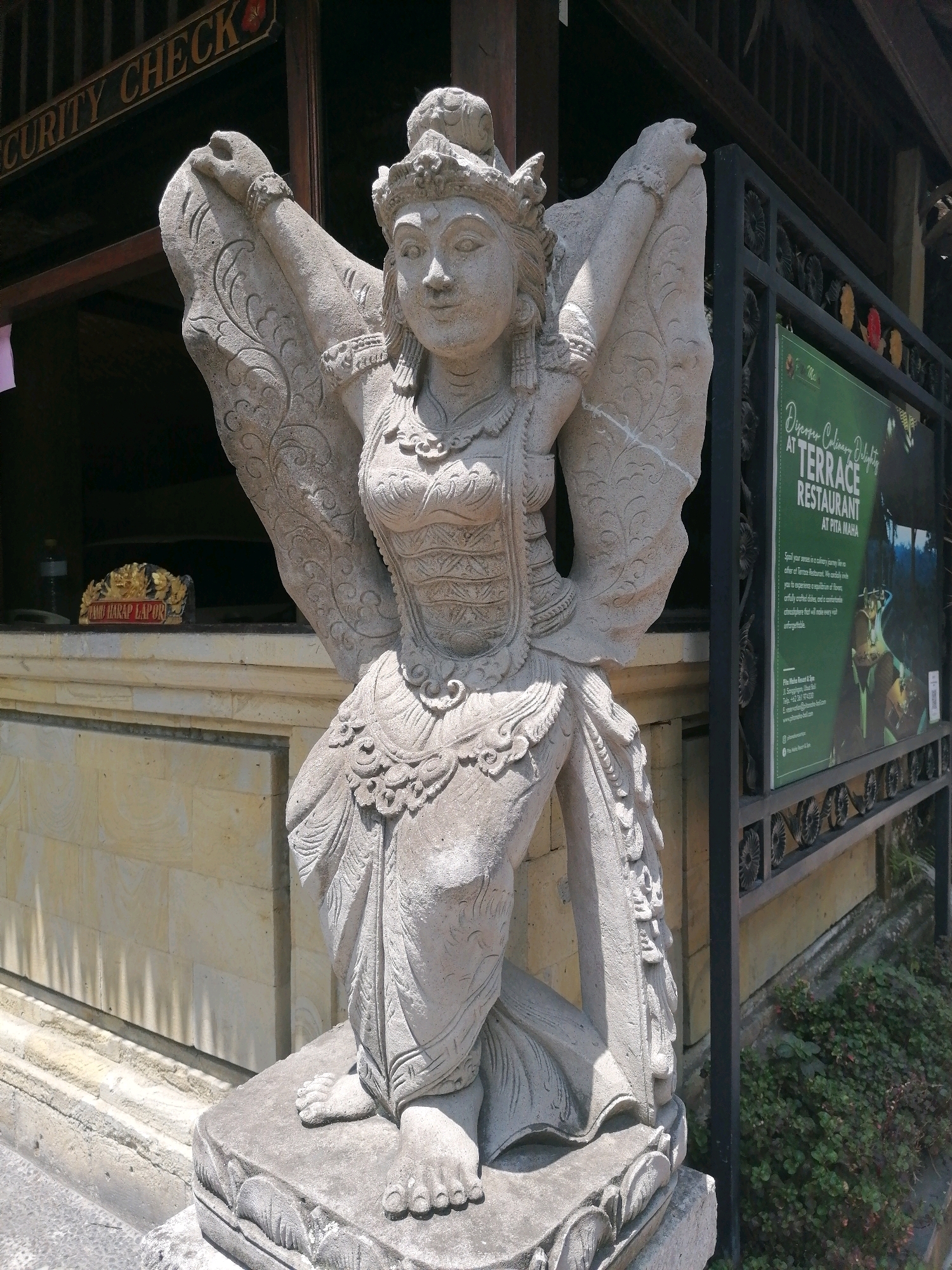 A stone statue of a mythical winged creature, likely a deity or demon from Balinese mythology, stands in front of a building with wooden architecture. The statue, carved from light-colored stone, depicts a humanoid figure with elaborate headdress, ornate clothing, and outstretched wings. The figure is in a kneeling pose, with its hands raised and touching its head. The background shows a building with a sign that reads "SECURITY CHECK" in English. To the right of the statue, a green sign advertises "TERRACE RESTAURANT AT PITA MAHA". The environment suggests a tropical location, with lush greenery visible in the lower right corner of the image. The overall scene depicts a cultural landmark likely located in the Ubud District, Indonesia, known for its rich artistic heritage and spiritual significance.