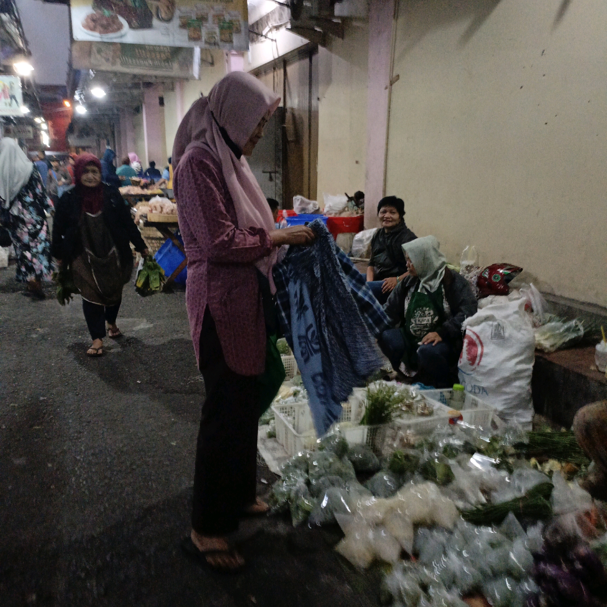 The image depicts a street market scene in Salatiga, Indonesia. In the foreground, a woman wearing a pink hijab and a patterned shirt is holding a blue and black checkered fabric. To her right, another woman in a green hijab and apron is seated, looking towards the viewer. Behind them, a woman in a dark jacket is also seated. Several white plastic bags containing produce are visible, along with a red bucket. In the background, more people and market stalls are present, with signs and lighting indicating a commercial area. The ground is paved, and shadows suggest it is either early morning or late evening.