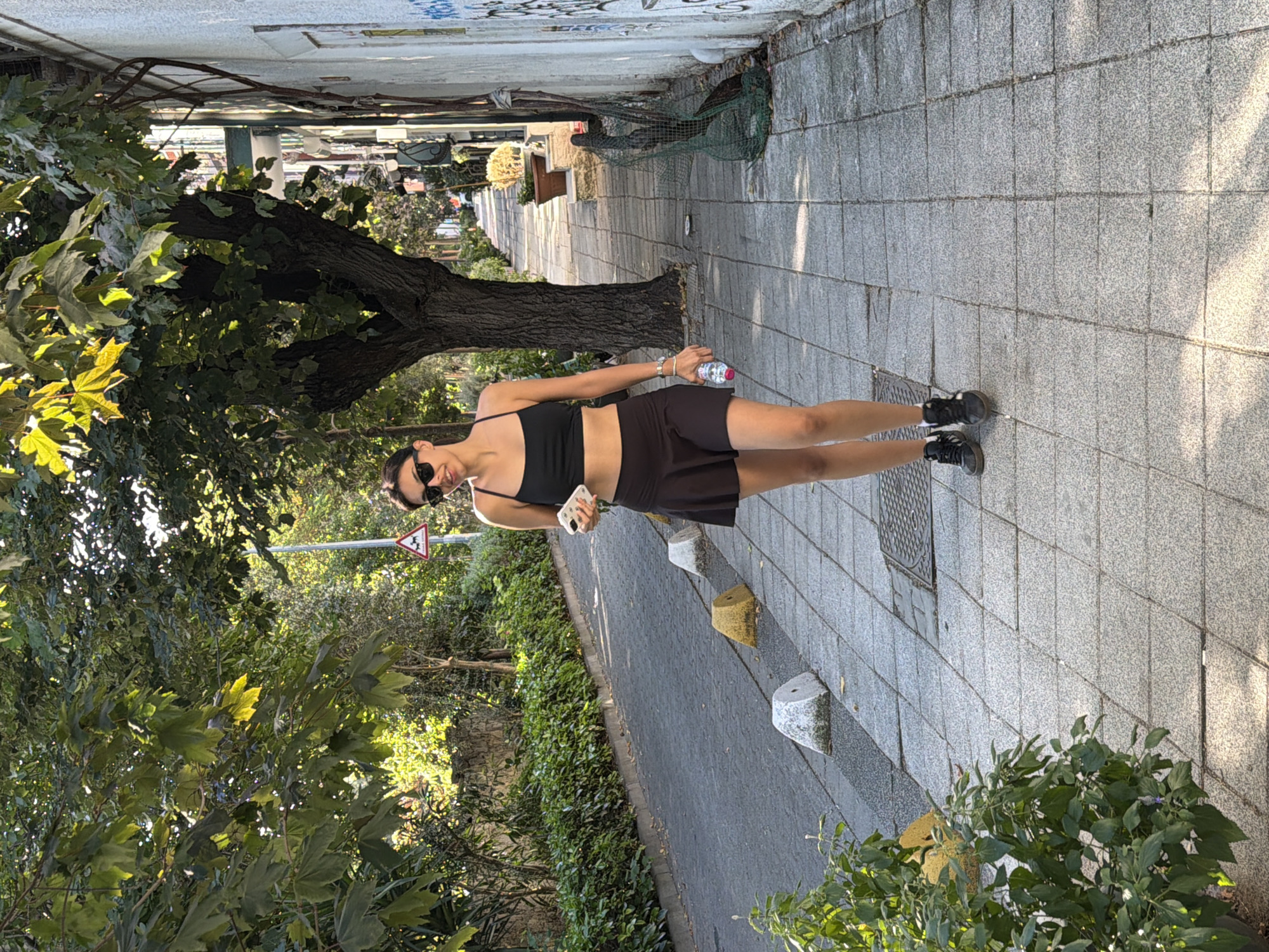 A young woman with dark hair and sunglasses walks along a tree-lined sidewalk in Kadıköy, Turkey. She is wearing a black crop top and a dark skirt, and is holding a smartphone in her left hand and a plastic water bottle in her right. The sidewalk is paved with grey tiles and there are several concrete bollards along the side of the street. The street is paved with grey asphalt. A traffic sign depicting a dog is visible in the background. The buildings on the right side of the street are painted white and feature graffiti. The scene is bathed in sunlight, casting shadows on the sidewalk and street. Lush green bushes line the left side of the street, and large trees with green leaves provide shade.