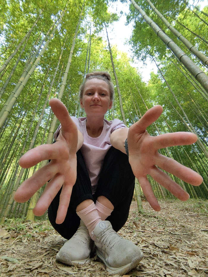 A fair-skinned individual is depicted in a low-angle, wide-angle shot within a dense bamboo forest in Mù Cang Chải, Vietnam. The subject, with light brown hair pulled into a bun, is squatting low to the ground and extends both arms forward towards the camera, presenting open hands with spread fingers, creating a forced perspective effect where the hands appear disproportionately large. The person wears a light pink short-sleeved t-shirt, dark-colored pants, light pink ankle socks, and light grey athletic shoes. A dark braided bracelet is visible on the left wrist. The background is dominated by numerous tall, slender green bamboo culms extending vertically upwards, with verdant foliage partially obscuring a bright sky. The foreground and ground beneath the subject are composed of earthy terrain covered with scattered dry leaves and debris.