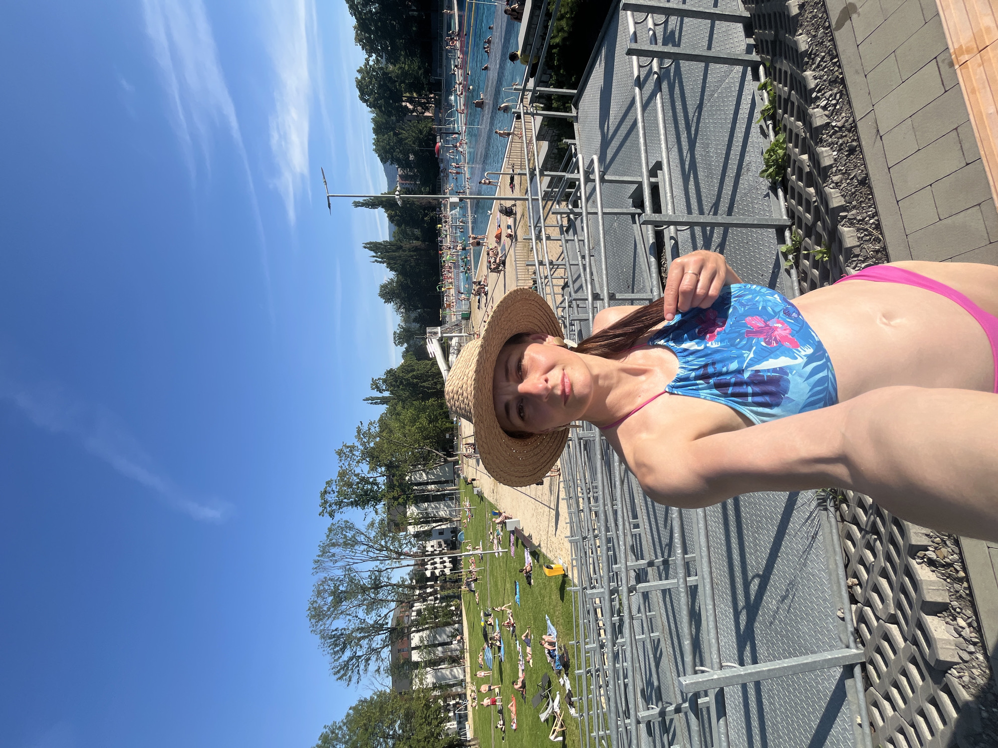 A woman wearing a straw hat and a blue and pink floral swimsuit takes a selfie outdoors at a swimming pool complex. She is standing in front of metal bleachers, with a large swimming pool and grassy areas with sunbathers visible in the background. The sky is clear blue with a few wispy clouds. The overall environment appears to be a recreational area on a sunny day.