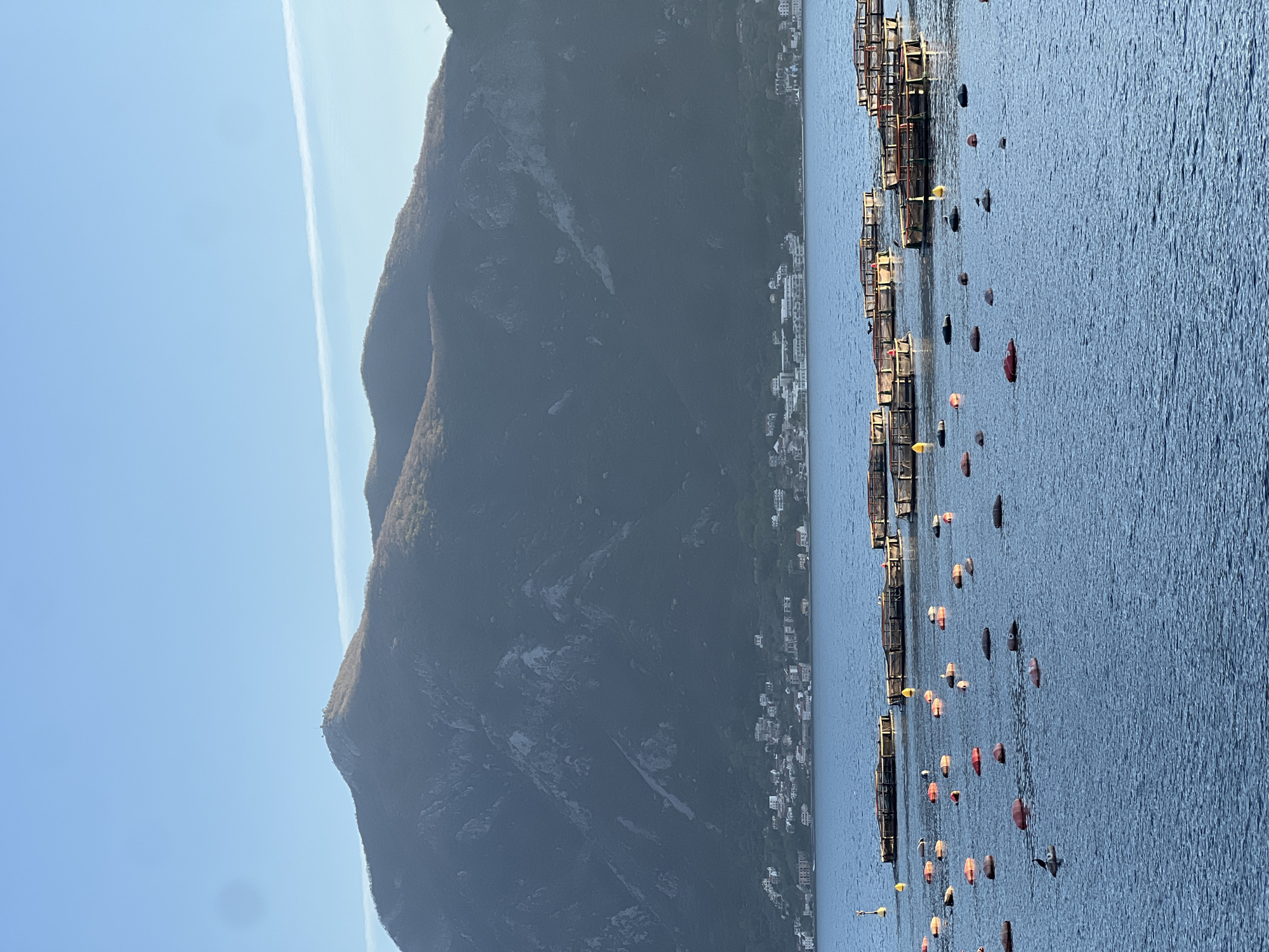 The image depicts a scene in Donji Orahovac, Montenegro. Visible are mountains in the background, with a town nestled along the shoreline. In the foreground is the Adriatic Sea, where floating fish farms marked by colorful buoys are seen. No people are clearly visible.