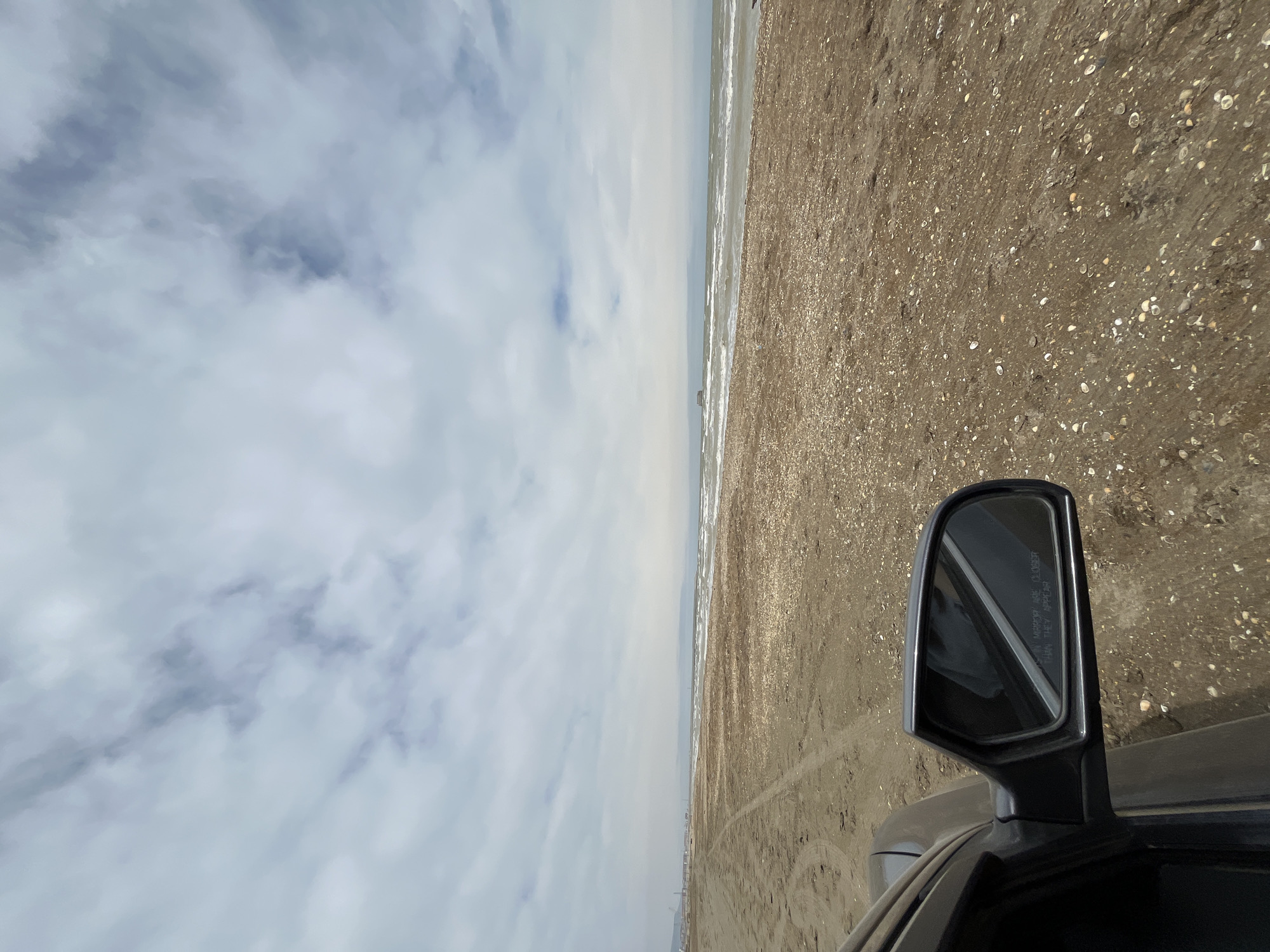 View of a beach in Kaspiysk, Russia, taken from inside a vehicle. The scene is divided roughly in half, with a cloudy sky to the left and the beach to the right. The beach is sandy and littered with shells, and stretches along the Caspian Sea. The side mirror of the vehicle is visible in the lower right corner of the frame. In the distance, a line of buildings can be seen along the shoreline.
