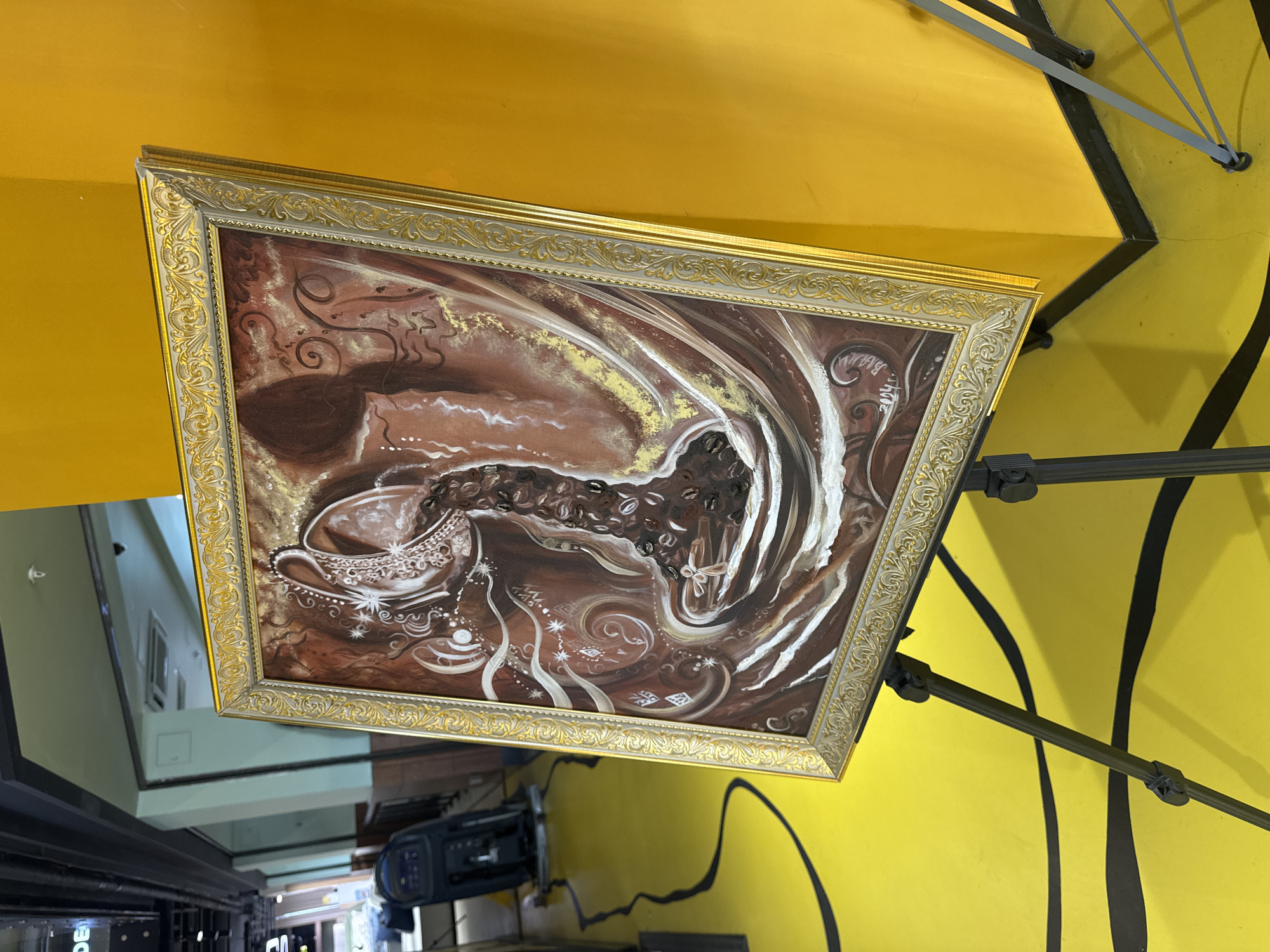 The image captures a framed painting hanging on a yellow wall. The painting features a coffee cup and coffee beans in abstract swirls and patterns, rendered in shades of brown, white, and gold. The frame is ornate with gold detailing. The wall is a bright yellow with thick black lines painted onto it. Metal support structures are visible, attached to the wall around the painting. The room appears to be part of a cafe or shop, based on the equipment visible in the reflection in the frame. The location is Moscow, Russia.