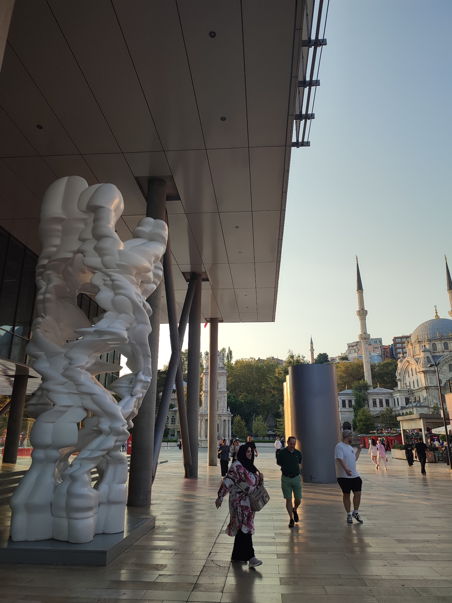 The image depicts an outdoor scene in the city of Beyoğlu, Turkey, likely during late afternoon or early evening, judging by the quality of light and long shadows. In the foreground, a large, abstract white sculpture with a fluid, organic form stands on a grey base. The sculpture is positioned in front of a modern building with a cantilevered roof supported by dark columns. The ground is paved with light-colored rectangular tiles, reflecting the sunlight.
Several people are visible in the midground and background. On the left, a woman wearing a headscarf and a patterned outfit walks towards the viewer, carrying a bag. To her right, two men walk away from the viewer. One man wears a green polo shirt and shorts, while the other wears a white t-shirt, shorts, and a cap. Further in the background, a group of people are walking in various directions, and some appear to be browsing at stalls or dining areas.
The background features a cityscape with traditional architecture. A mosque with a large dome and tall minarets is prominently visible, nestled amongst trees and other buildings on a hillside. The sky is a clear, pale blue. The overall impression is a blend of contemporary urban design and historical cultural landmarks.