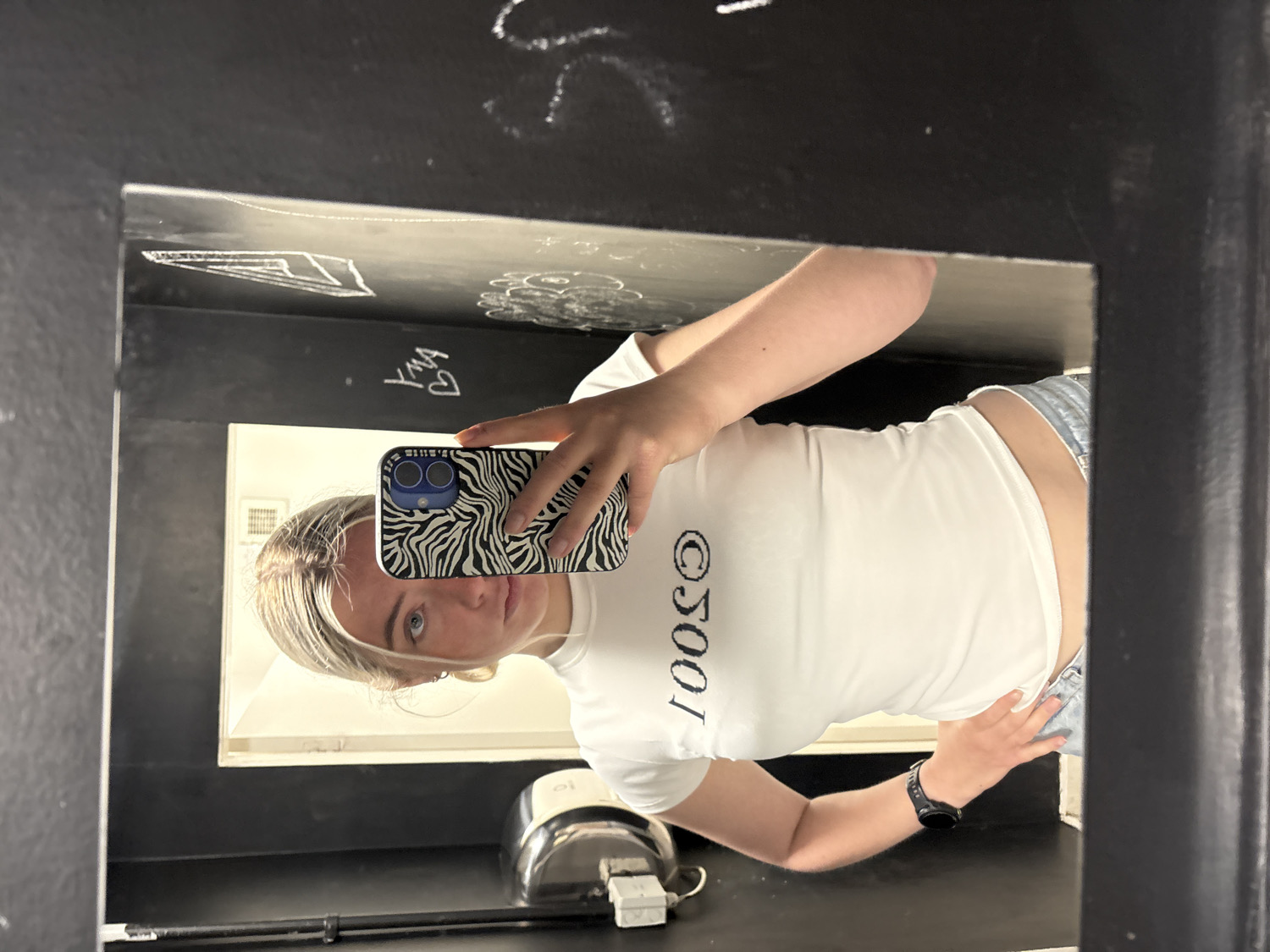 The image, rotated 90 degrees clockwise, depicts a young woman taking a mirror selfie indoors. She is positioned in front of a mirror with dark, possibly black, walls that are covered in white graffiti and chalk drawings, including "YMA" with a heart symbol.
The woman has blonde hair and blue eyes, and she is wearing a white short-sleeved top and light-wash denim shorts or jeans. A black smartwatch is visible on her left wrist. She holds a smartphone with a distinctive black and white zebra print case to take the picture, partially obscuring her face. The word "1001" is printed vertically in black letters on the front of her white top.
The setting appears to be a restroom or a changing area, with a sink fixture partially visible in the reflection behind her. The woman maintains a neutral expression for the photo. While the location context is provided as Manchester, United Kingdom, there are no specific visual cues within the image itself to confirm this. The lighting is artificial, suggesting an indoor environment at an indeterminate time of day.
