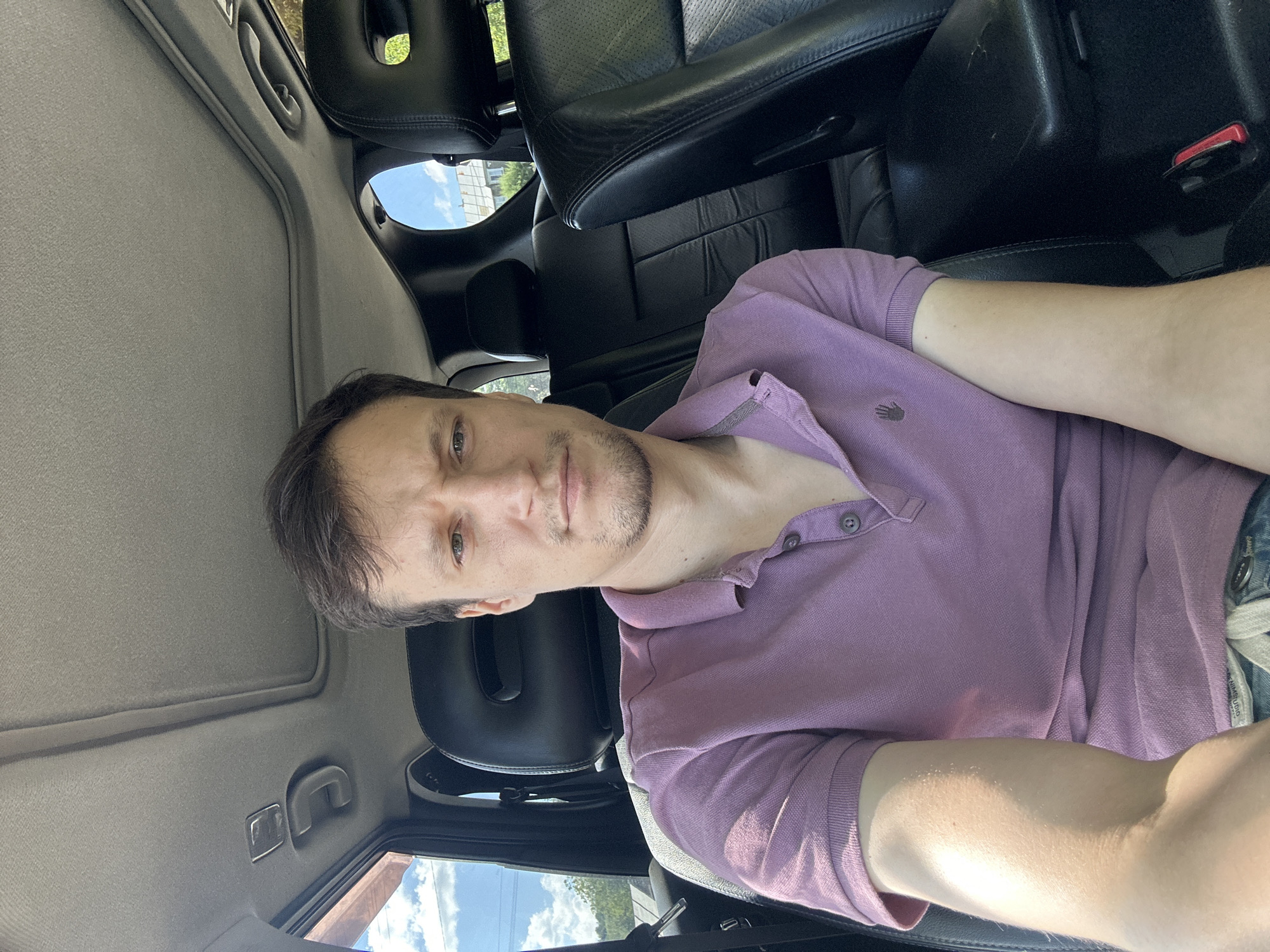 A young man with short brown hair and a slight stubble is seated in the passenger seat of a car. He is wearing a purple polo shirt. The interior of the car is visible, including black leather seats and a sunroof. Outside the car, green trees and a blue sky with white clouds can be seen. The image appears to be taken during the daytime. There is no visual information within the image that specifically indicates the location as Makiv, Ukraine.