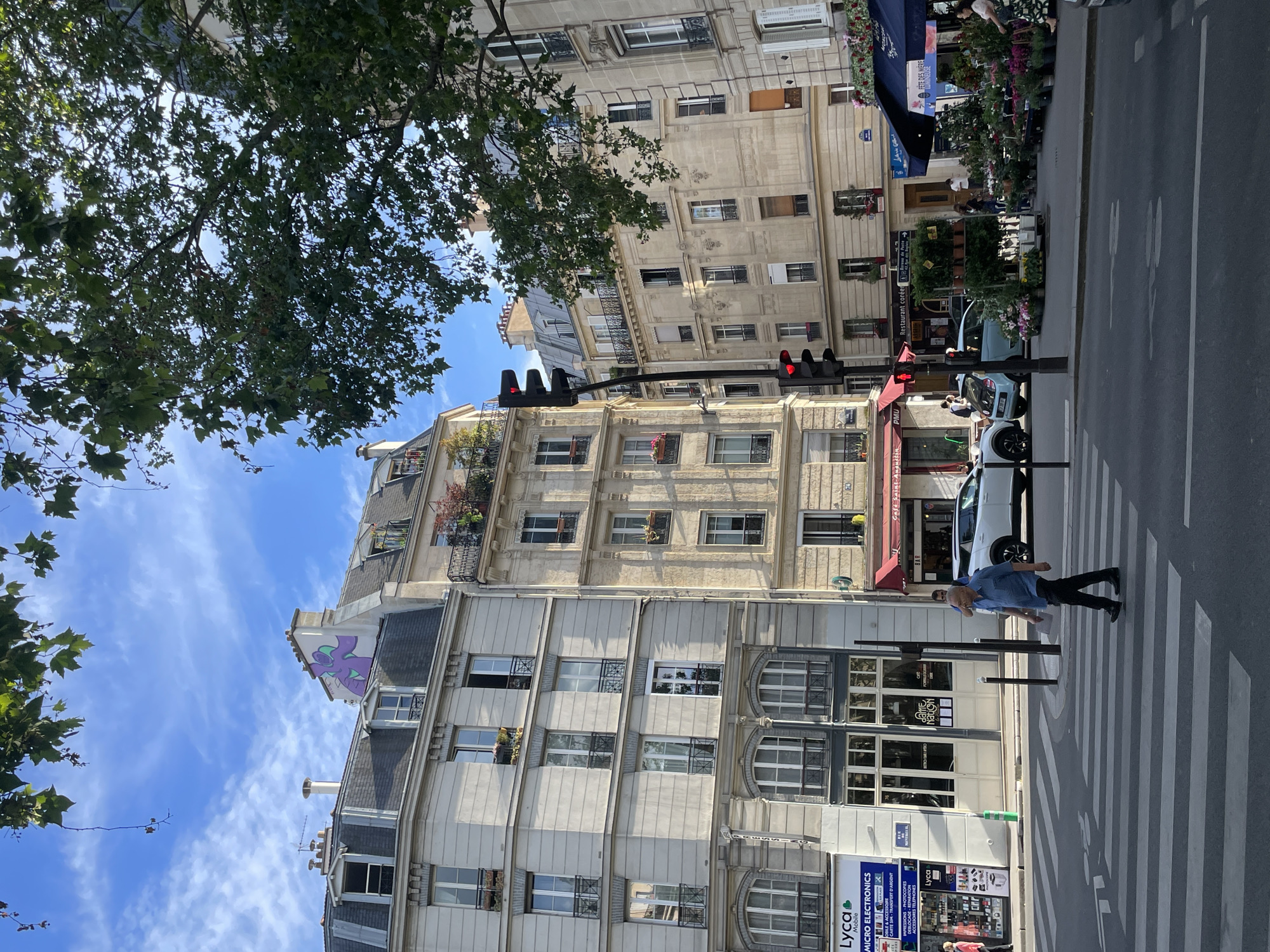 This vertical image depicts a bustling street scene in Paris, France, under a clear, sunny sky with scattered wispy clouds. Lush green tree foliage partially frames the top left of the frame, indicating a tree-lined street.
The scene is dominated by classic Parisian architecture, featuring multi-story Haussmannian buildings with light-colored stone facades, intricate detailing, and numerous windows, some adorned with small balconies and flower boxes. On the upper gable of one building, a striking purple and green abstract mural is visible.
In the foreground, a pedestrian wearing a blue shirt and dark pants is prominently walking across a zebra-striped crosswalk. Traffic lights displaying red are visible, positioned above the street. Several parked cars, including a light blue compact car, are seen along the curb.
The ground floors of the buildings house various businesses. On the right side, a vibrant flower shop displays an abundance of colorful plants and flowers, with several individuals visible near the storefront, including a sign reading "FETE DES MERE" (Mother's Day). Other establishments include a "Restaurant coréen" (Korean restaurant), a "BAR," and a "PMU." A shop named "MICRO ELECTRONICS" advertises "Lyca Mobile," mobile & accessories, SIM cards, and money transfer services. The overall atmosphere suggests a typical daytime urban environment with commercial activity and pedestrian movement.
