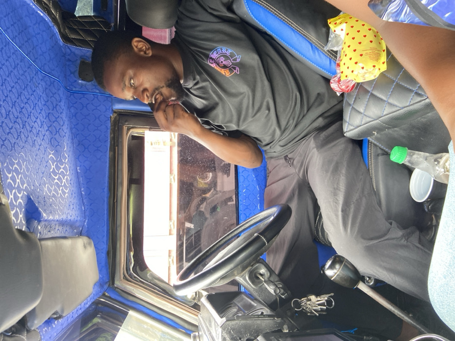 An adult man in a black shirt sits in the driver's seat of a vehicle. The interior features a blue patterned wall and diamond-stitched upholstery. The man looks down and has a finger near his mouth. A steering wheel, gear shift, and keys are visible. A yellow bag with red hearts and a discarded plastic bottle sit on the passenger seat.