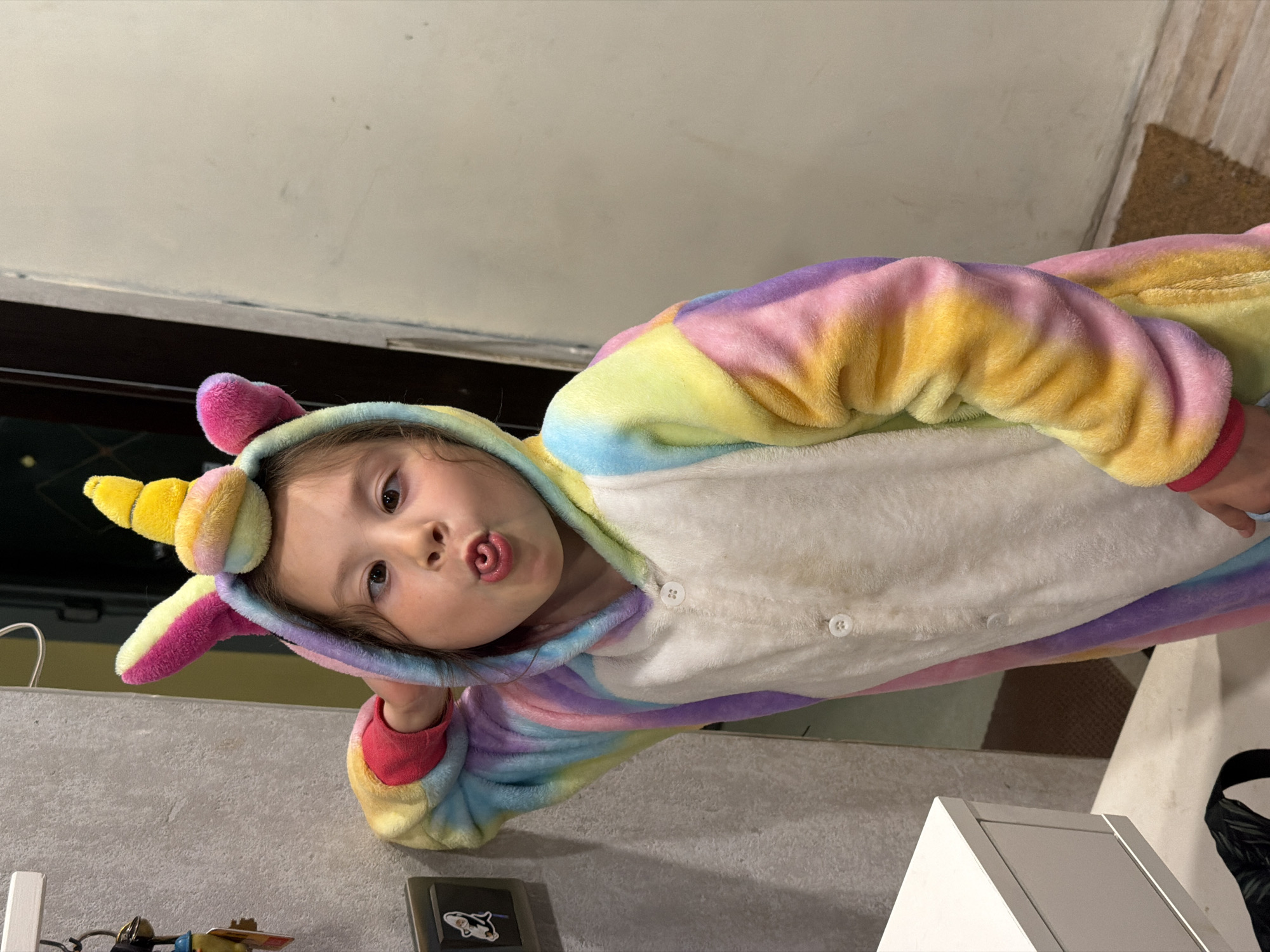 A child in a pastel rainbow unicorn onesie with a yellow horn and pink ears is sticking their tongue out. The child appears to be indoors. A mat and a white cabinet are partially visible.
There is no evidence indicating this image was taken in Perm, Russia.