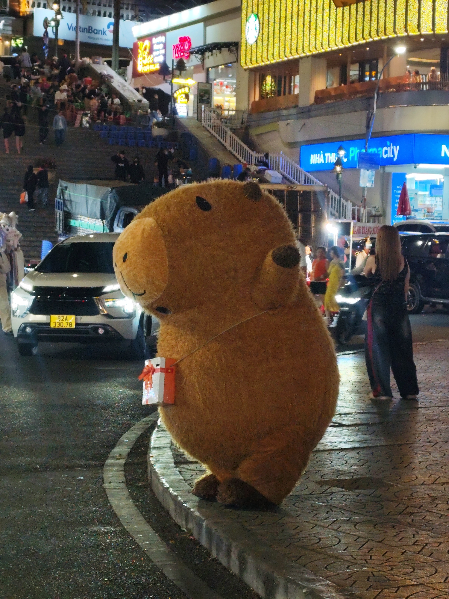 A fuzzy brown mascot figure wearing a small gift stands on a patterned sidewalk beside a curb and street in Da Lat, Vietnam. The street shows some water and streetlights in the background illuminate the scene. A white SUV with a license plate "92A 330.78" is parked on the street. A woman with long blonde hair wearing a black top and patterned pants stands on the sidewalk to the mascot's right. Other people stand around in the background, along with other vehicles. Above the street, stairs lead up to a crowd of people watching something on the steps, and a building in the background has blue "NHA THUOC Pharmacy" signs. The architecture of the area suggests an urban setting with businesses open at night.