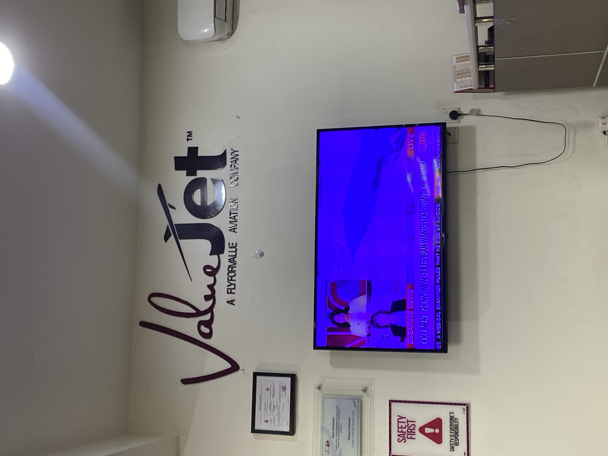 The image shows the interior of what appears to be an office or reception area. On the wall is the logo of a company called "Value Jet: A Fly for Value Aviation Company." Below the logo are three framed items: a certificate, a "Vision and Mission Statement," and a "Safety First" sign with a red triangle and exclamation point. A television is mounted on the wall, displaying "Breaking News" from CNN. There is no evidence in the photograph to confirm whether the image was taken in Ikeja, Nigeria.