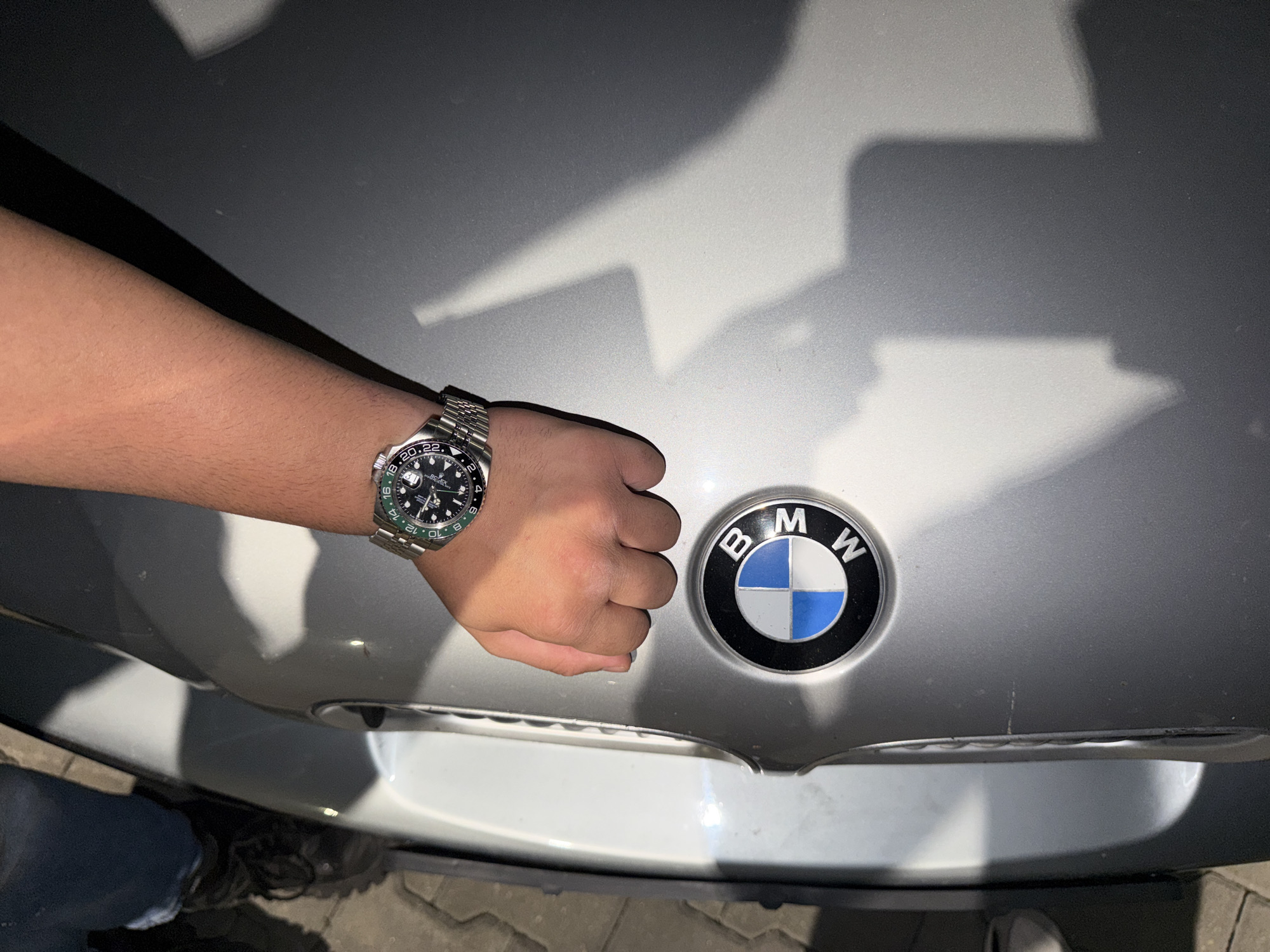 The image captures a person's arm, adorned with a luxury watch, resting on the hood of a car. The primary subjects are a person's right arm and closed fist, wearing a stainless steel Rolex GMT-Master II "Sprite" watch with a green and black bezel and a Jubilee bracelet. The watch dial clearly shows "ROLEX" and partially "GMT-MASTER II", along with numbers on the bezel. Adjacent to the fist is the circular BMW emblem, prominently displayed on the silver or light grey car hood. The "B M W" lettering is visible within the emblem.
The scene appears to be outdoors, with distinct, sharp shadows cast across the car's surface, suggesting strong lighting conditions, possibly direct sunlight or powerful artificial light. A paved surface, resembling cobblestones, is visible at the bottom of the frame, hinting at a parking lot or driveway setting. Part of a person's leg, clad in blue jeans and shoes, is also visible in the lower-left corner. The activity depicts the person posing their arm and watch on the car, likely showcasing both items. No specific emotions are discernible.