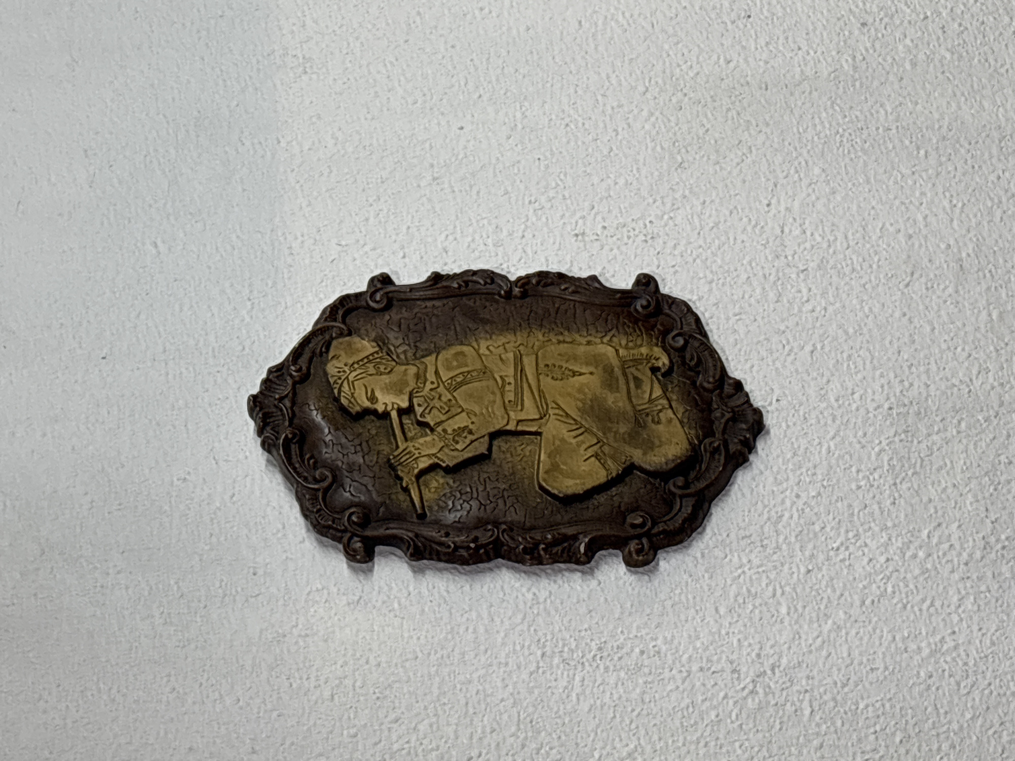 The image shows a bronze-colored plaque affixed to a white wall. The plaque depicts a figure, possibly a soldier or historical figure, kneeling and holding a long, slender object, possibly a weapon or staff. The plaque has an ornate, oval-shaped border with decorative scrolls and embellishments. The background wall appears to be textured and painted white.
Given the historical context of the figure depicted, it is likely that the plaque is located in Pleven, Bulgaria, possibly within a historical museum, memorial site, or public building. The figure, his attire, and the staff represent a historical person from the time period of the siege of Pleven in 1877-1878 during the Russo-Turkish War.
