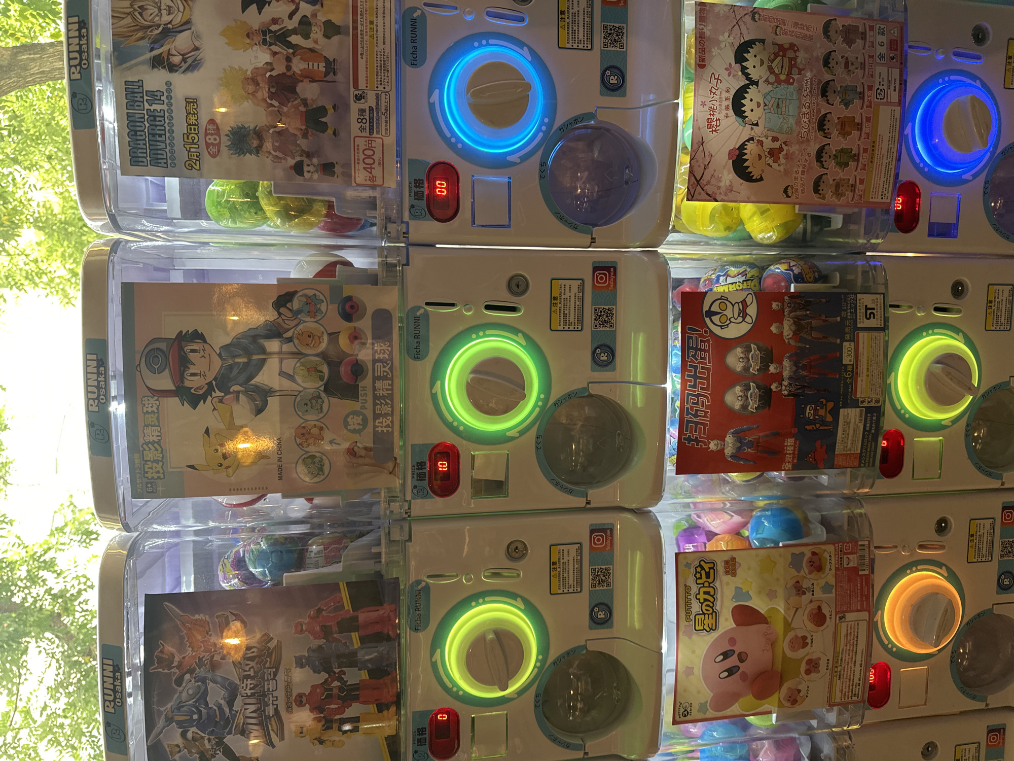 The image shows a wall of "Gashapon" or capsule toy vending machines. Each machine displays different anime and game character merchandise. The characters include "Dragon Ball," "Pokemon," "Kirby," and "Chibi Maruko Chan." The machines are white with colorful circular light-up displays, coin slots, and prize retrieval chutes. Some machines are priced at 400 yen, others at 300 yen. The labels are in Japanese. The background includes a tree and suggests an outdoor setting. The location is likely not in Madrid, Spain.