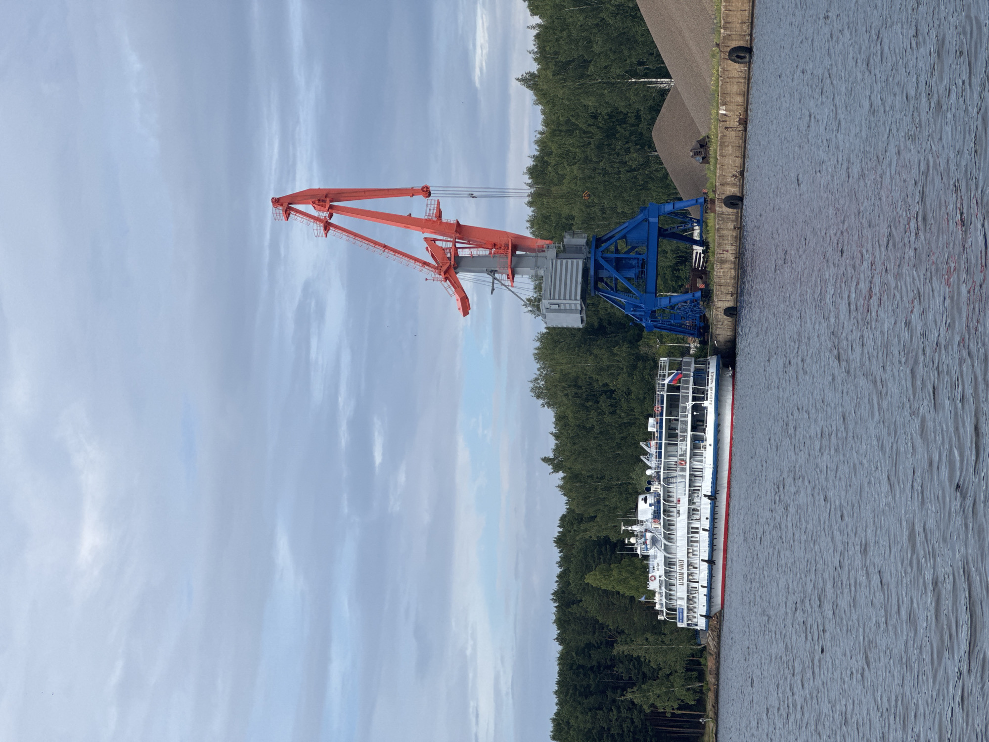 The image depicts a serene, overcast day at a port in Chaikovsky, Russia. A large, red and blue industrial crane dominates the left side of the frame, its imposing structure contrasting with the natural beauty of the surrounding environment. To the right of the crane, a white passenger ship is docked along the concrete embankment. The ship, with multiple decks and railings, appears ready for departure or has recently arrived. The water in the foreground is calm, reflecting the muted colors of the sky and surrounding trees.
The backdrop features a dense, green forest, suggesting a natural landscape bordering the industrial port. The sky is filled with soft, wispy clouds, indicating a mild, possibly cool, day. The overall atmosphere is one of quiet industry and readiness. There is no visible activity of people, but the presence of the ship and crane implies ongoing port operations.
A piece of text is visible on the side of the ship, but it is partially obscured and difficult to read in its entirety. However, the letters "ЧАЙКОВСКИЙ" (Chaikovsky) and "БАЛАГАН" are discernible, confirming the location. The time of day appears to be either morning or late afternoon, given the diffused light. The scene evokes a sense of calm, with the powerful machinery of the crane and the sleek design of the ship harmonizing with the tranquil natural setting.
