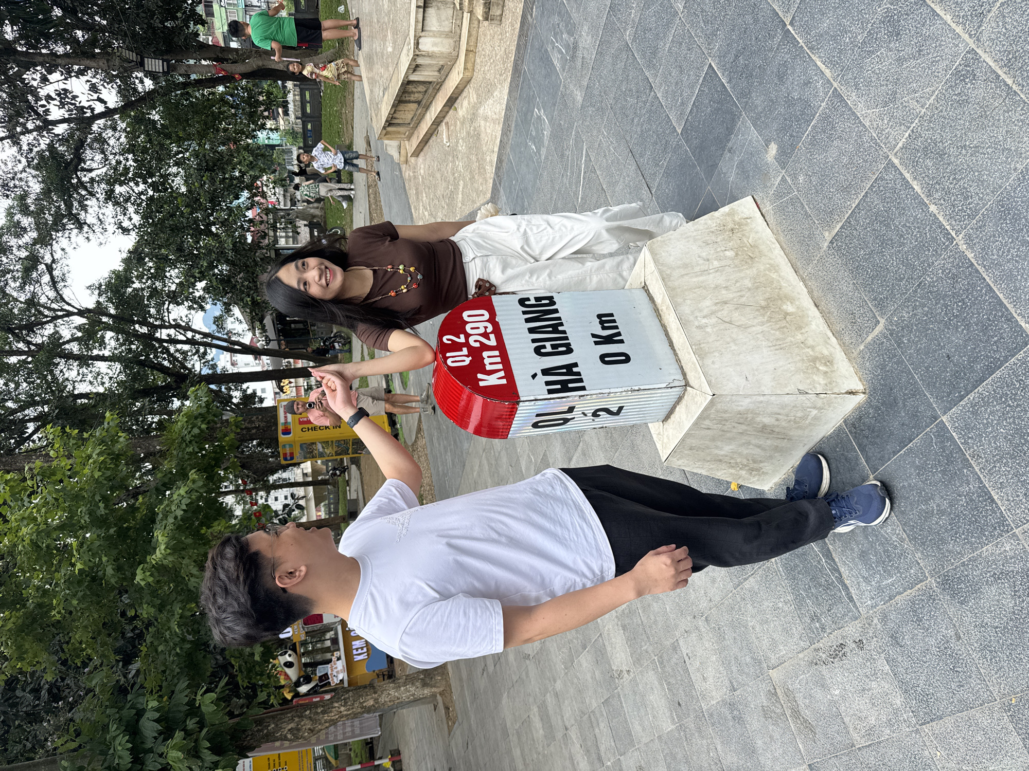 A medium shot captures a man and a woman interacting with a distance marker in Hà Giang 1 Ward, Vietnam. The man, dressed in a white t-shirt and black pants, reaches out to touch the woman’s hand. She is lying on the distance marker, wearing a brown top and white pants. The marker indicates "Hà Giang 0 Km" and "QL 2 Km 290". The surroundings include trees and other people in the background.