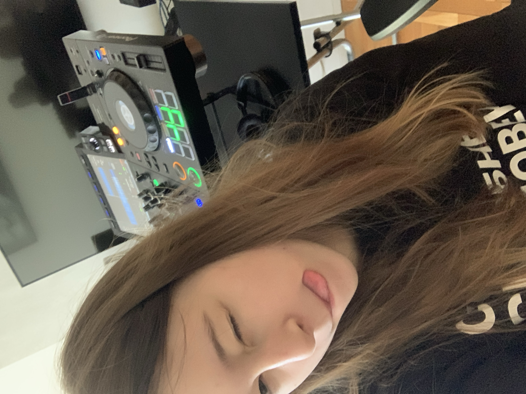 A young person with long brown hair is positioned close to the camera, sticking out their tongue and winking. Behind them, there is a DJ mixer on a black table, with a pair of headphones next to it. A monitor is visible in the background. No information available to identify location as Vozdovac Urban Municipality, Serbia.
