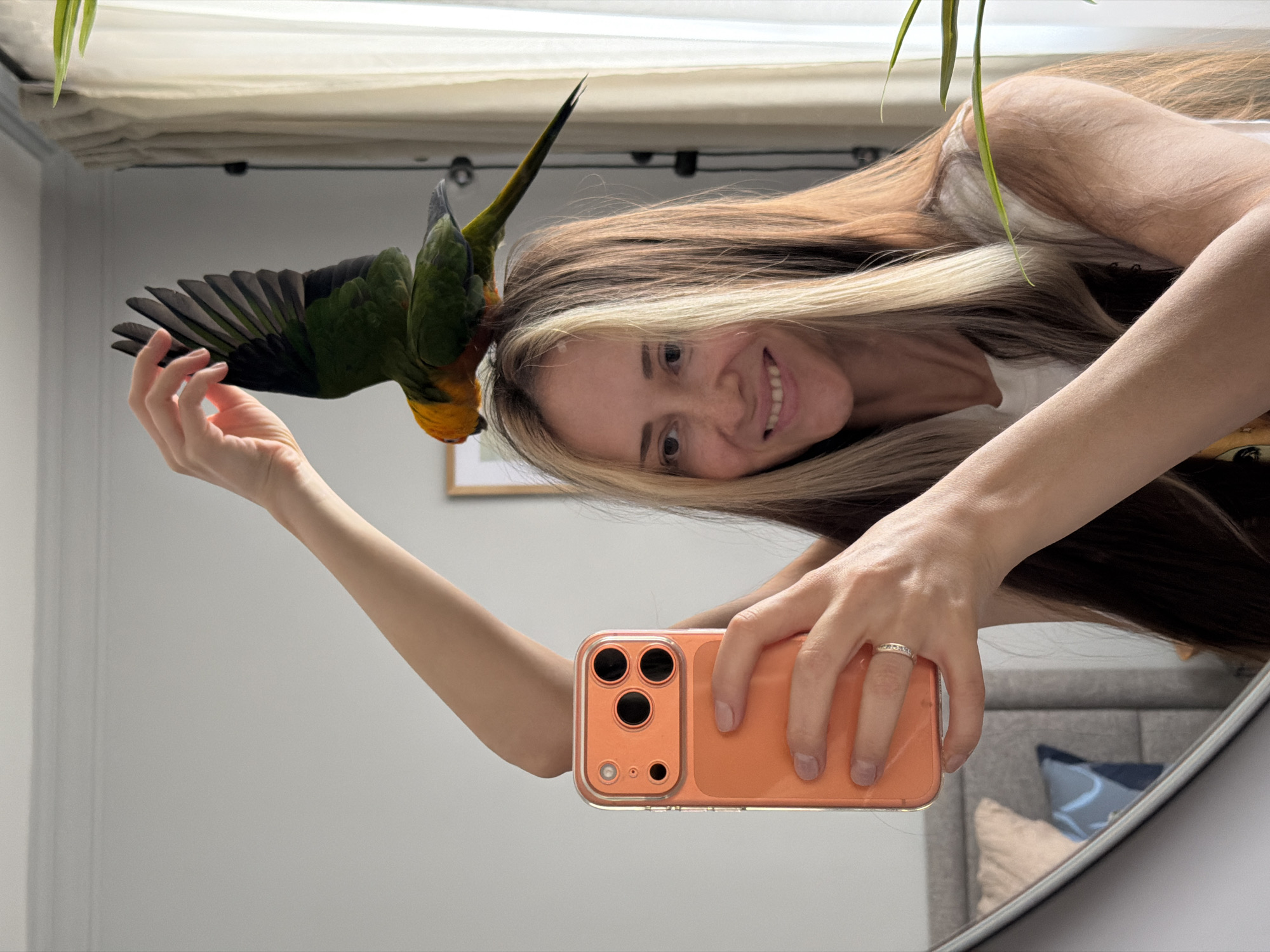 A fair-skinned woman with long brown hair and blonde highlights takes a selfie in a mirror with her orange-colored phone, and a green and orange conure parrot is perched on her head. The woman smiles at her reflection. The reflection shows a headboard with pillows behind her. The wall is light gray, and a framed picture hangs on the wall behind the parrot. There is a window with a blind at the top of the frame. The presence of a plant hanging down from the top left corner of the image suggests that this is an indoor scene. While the image lacks location data, it is tagged as being taken in Khimki, Russia.