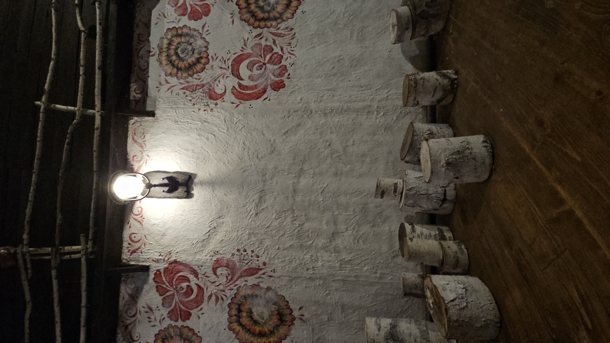 The image depicts an interior scene with a textured white wall adorned with red and yellow floral paintings in a folk art style. A single light fixture hangs from the ceiling, illuminating the wall. Below the wall, a collection of birch logs of varying sizes are arranged on a dark wooden surface. The ceiling features exposed wooden beams and what appear to be thin branches or twigs. The overall impression is rustic and decorative. This scene is located in a restaurant in Ulyanovsk, Russia.