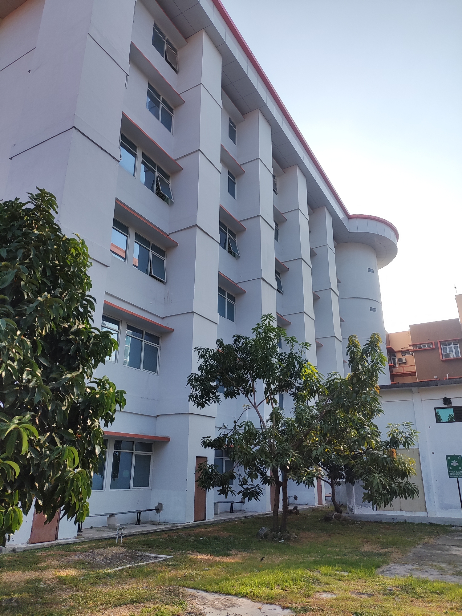 The image depicts a modern, multi-story building with a white facade and orange trim around its numerous windows. The building appears to be a public or institutional structure. In the foreground, several trees with green foliage are partially obscuring the lower floors of the building. The ground is covered with grass, and there are some paved pathways and benches visible. A sign with text in what appears to be Indonesian is present on the right side of the image, along with a green trash receptacle. The sky is clear and bright, suggesting a daytime scene.
Based on the presence of the Indonesian language sign, the location can be identified as Cirebon, Indonesia. The building itself is likely part of an educational institution or a government facility within the city. There are no people visible in the image, and the scene is static. The overall impression is one of an institutional setting, likely well-maintained and situated in a pleasant, green environment.