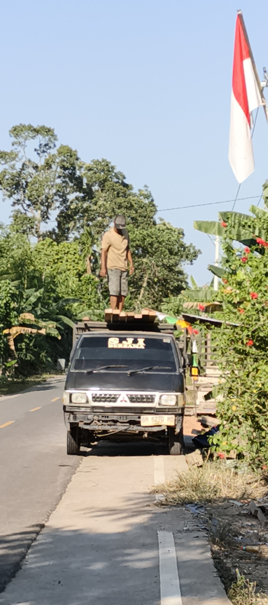 A man wearing a light brown t-shirt and grey shorts stands on top of a loaded truck. The truck is black and parked on the side of a road. The man is standing on wooden planks that are stacked on the truck. The background is filled with lush green trees and vegetation. A flag, red and white, is visible in the upper right corner of the image. The license plate of the truck reads "BH 6270 HD". The scene appears to be in Muaro Jambi, Indonesia, likely along a rural road.