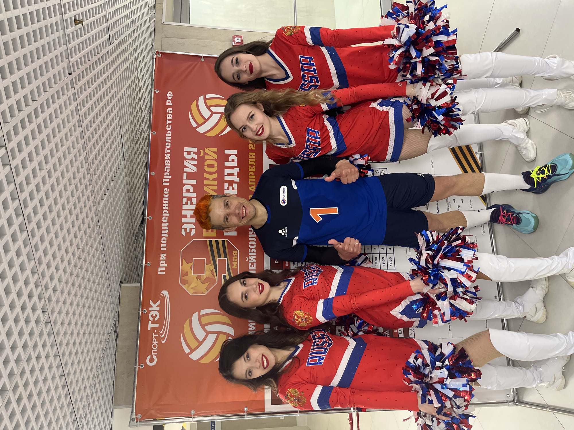 Here is a description of the image:
**Scene**: A group photo featuring four women in cheerleader outfits and a man in a blue and black athletic uniform with the number 1, likely a volleyball player. They are posing in front of a banner with text in Russian and a volleyball graphic.
**People**:
* **Cheerleaders**: All four women are wearing matching red, white, and blue cheerleading uniforms, and white boots. They are holding pom-poms in the same colors. All of them are smiling.
* **Volleyball Player**: The man is wearing athletic gear and has an orange tint to his hair. He is giving a thumbs-up gesture with both hands.
**Objects**:
* **Banner**: The banner is mostly red and contains logos, Russian text.
* **Pom-Poms**: Red, white, and blue
* **Volleyball**: The banner includes graphics of a volleyball.
* **Boots**: The cheerleaders have white boots.
* **Athletic Shoes**: The volleyball player has a pair of running sneakers in different colors.
**Environment**: The setting appears to be indoors, potentially inside a sports venue. A white textured wall is visible in the background.
**Action and Interactions**: The group is posing for a photograph, as all members are smiling and interacting with the camera. The cheerleaders and volleyball player are positioned next to one another.