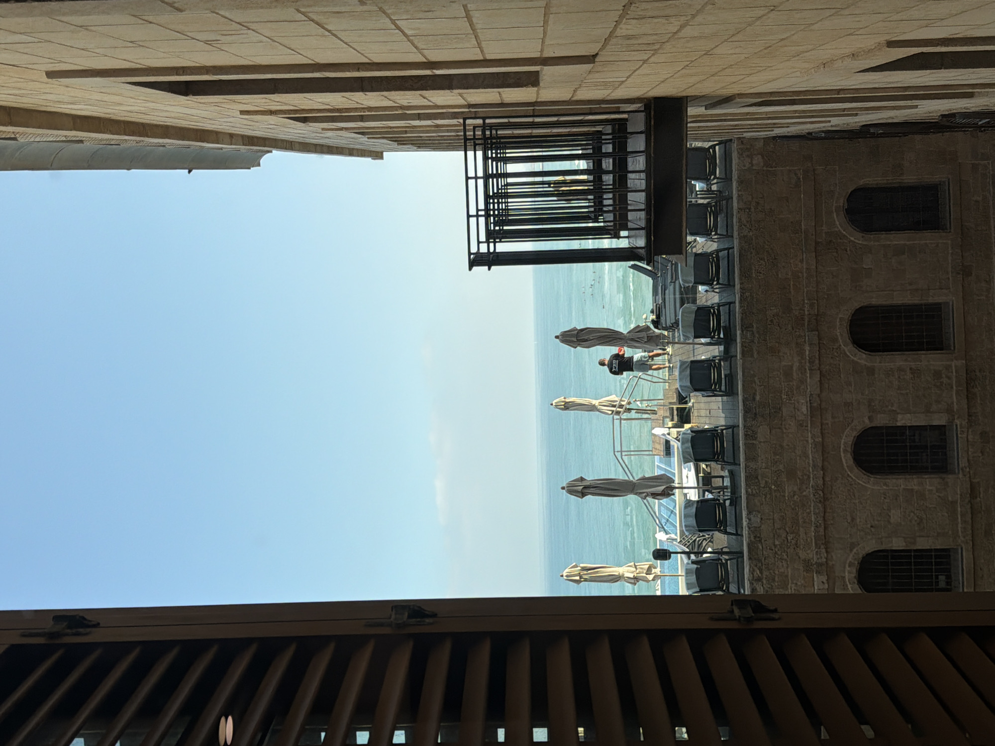 The image, when viewed in its correct upright orientation, captures a serene coastal scene in Tel-Aviv, Israel, observed from a narrow passage between two stone buildings. The composition is framed by light-colored stone architecture: a building on the right with two prominent arched windows featuring grilles, and another forming the left side of the passage.
In the mid-ground, an outdoor seating area or terrace, likely part of a cafe or restaurant, extends towards the sea. This area is furnished with numerous dark chairs, some arranged around tables, and several large, light-beige or grey patio umbrellas, mostly in a closed or partially closed state. A black metal structure, resembling a balcony or a fire escape, juts out from the left building above the terrace.
One individual is clearly visible on the terrace, standing and facing the sea. This person is wearing a dark t-shirt with white Hebrew text (partially discernible as possibly "בֶּקֶר" - morning, or a similar word), shorts, and a cap. Beyond the terrace, the calm, turquoise waters of the Mediterranean Sea stretch out under a clear, bright blue sky, meeting the horizon.
In the immediate foreground, a dark wooden slatted structure, possibly a shutter or part of a window frame, is visible, complete with a metal latch. The bright, even lighting suggests either a clear morning or late afternoon. The overall scene conveys a sense of tranquility and leisure, characteristic of a seaside promenade or a cafe overlooking the historic Jaffa Port.
