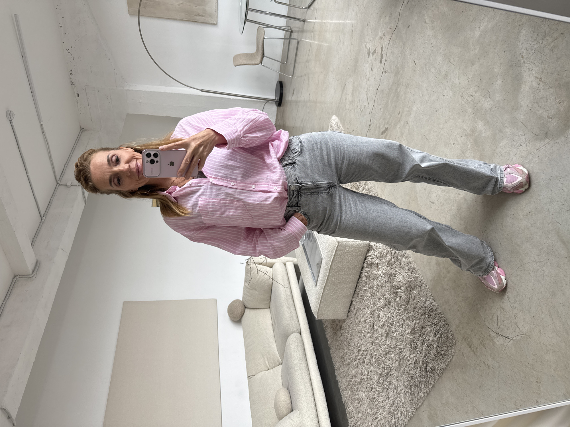 A woman is taking a selfie in a room with modern decor. She is wearing a pink striped button-down shirt, grey jeans, and pink and white sneakers. She is holding a pink phone in her right hand, taking the picture. The room has a beige sofa, a textured rug, and a polished concrete floor. There's also a metal floor lamp and dining table with chairs in the background. A large, framed piece of art leans against the wall. The location is not indicated in the image.