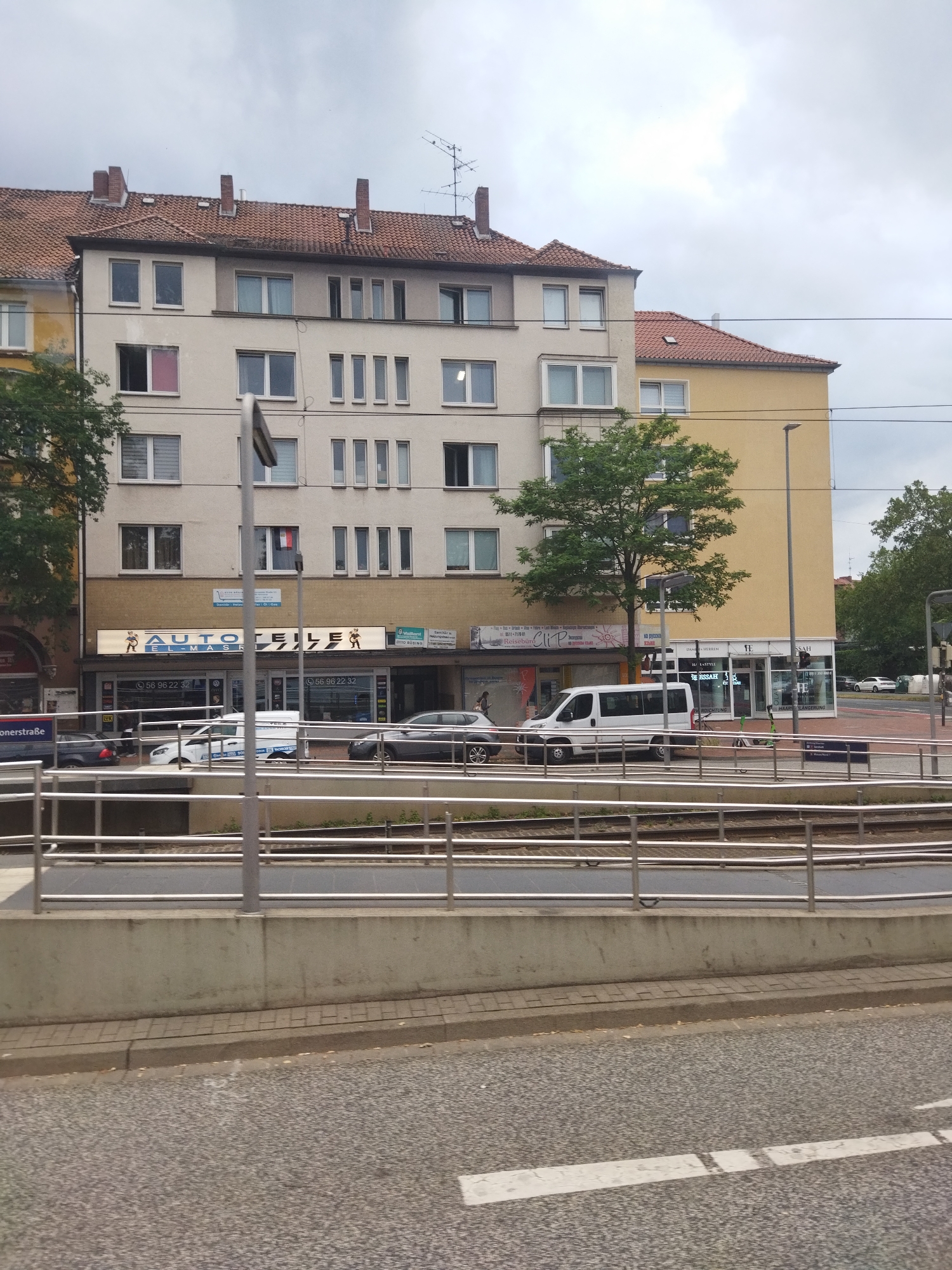 The image shows a street view in Hanover, Germany, with a multi-story building on the right side of the frame. The building has a light-colored facade and a red tiled roof. There are several windows on the upper floors. The ground floor of the building appears to house commercial establishments, including a business with a prominent sign that reads "AUTO TEILE". Several vehicles, including a white van and cars, are parked in front of the businesses. A street and tram tracks are visible in the foreground, separated from the building by a metal railing and a concrete barrier. The sky is overcast, suggesting a cloudy day. The image appears to be taken from a moving vehicle, possibly a tram or car, looking towards the buildings across the street.