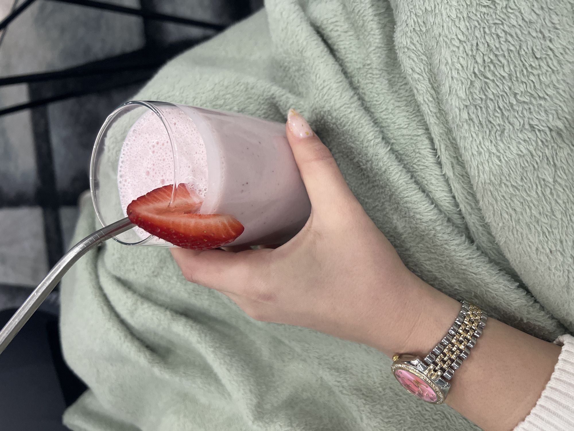 A person holds a glass of pink smoothie with a strawberry slice attached to the rim by a silver straw. The person wears a two-tone metal watch with a pink face on their left wrist and is covered by a light green, plush blanket. The background is blurry, showing a black and white pattern. The image's location is unknown and cannot be determined as Yambol, Bulgaria.