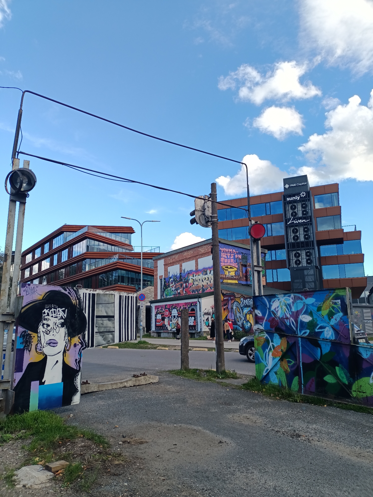 The image captures a street scene in Tallinn, Estonia, characterized by vibrant street art and modern architecture. In the foreground, a prominent mural depicts a stylized portrait of a woman wearing a wide-brimmed hat, rendered in black and white with accents of purple, yellow, and blue. To the right of this mural, another section of graffiti displays colorful floral motifs.
The street itself is paved and appears to be a road or pathway, with a concrete barrier and patches of grass on either side. A traffic signal pole with a red light is visible, along with power lines crisscrossing the sky.
In the background, a multi-story building with a distinctive stepped design, featuring a dark-colored facade and large glass windows, dominates the scene. Adjacent to this structure is a brick building adorned with further graffiti, including cartoonish figures and a "STOP" sign. A tall, dark tower-like structure is mounted on the modern building, displaying the logos "P&E Capital" and "sunly" with the word "Liven" beneath it.
A few individuals are present in the middle ground, near the brick building, and a dark-colored car is seen driving on the road. The sky is a bright blue with scattered white clouds, indicating fair weather. The overall impression is a blend of urban decay and artistic renewal, with street art juxtaposed against contemporary architectural elements.