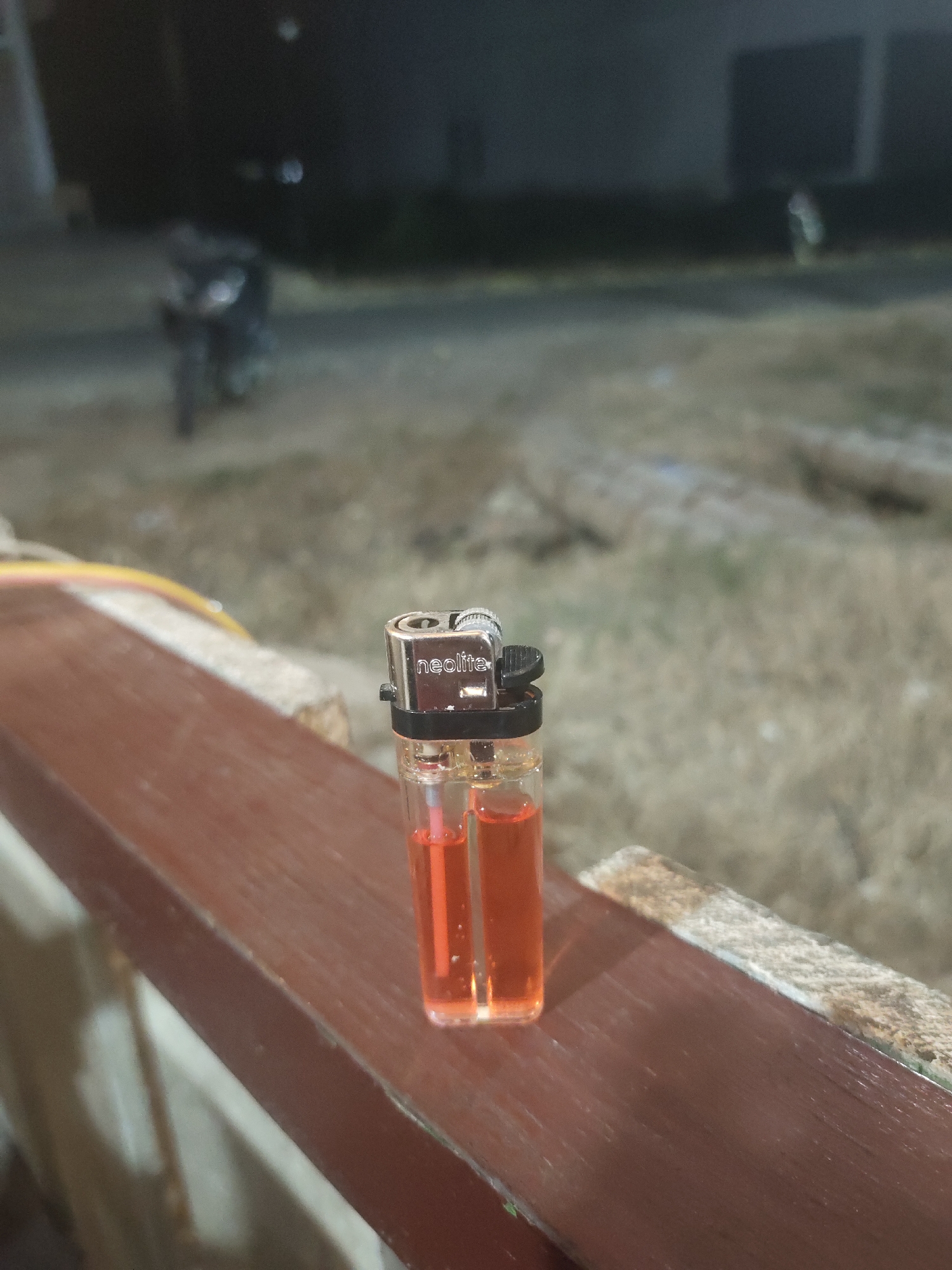 A clear plastic lighter with orange fluid rests on a brown ledge. The word "neolite" is visible on the lighter's metal top. In the blurred background is a motorcycle, foliage and buildings. The image was taken at night, likely in Keudematangkuli, Indonesia.