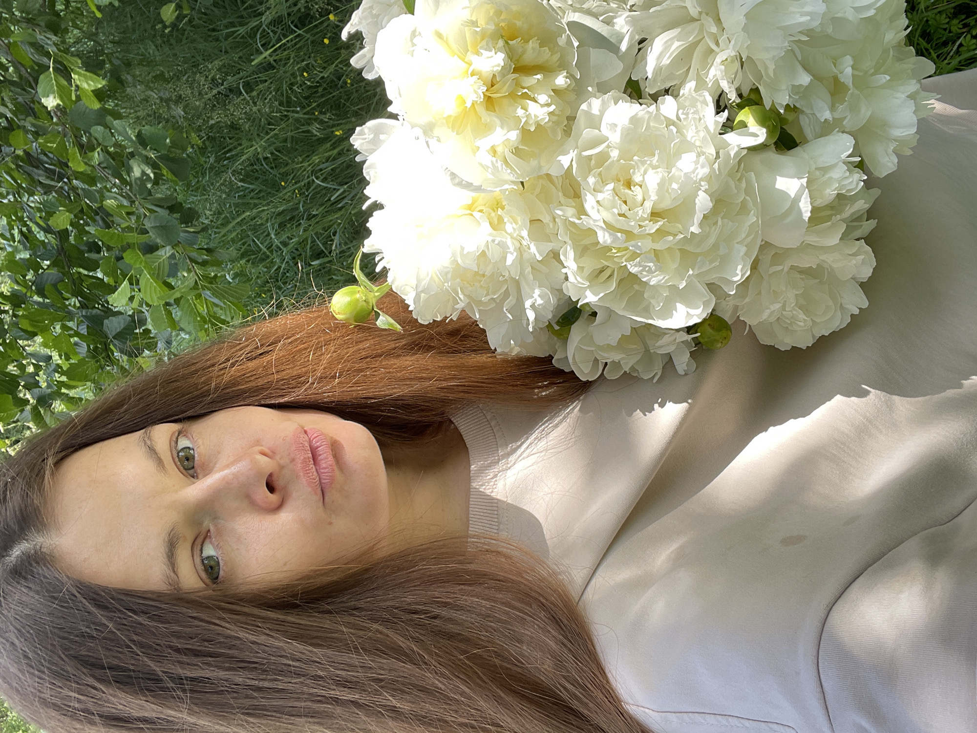 This is a portrait of a young woman with long, brown hair, lying outdoors. She is holding a large bouquet of white peonies. The woman's face is turned towards the camera, and she has green eyes and light-colored lips. She is wearing a light-colored, long-sleeved shirt.
The background consists of lush green grass and trees, suggesting a natural, outdoor setting. Sunlight filters through the leaves, casting dappled shadows on the woman and the flowers. The overall mood is serene and natural. The presence of flowers and the outdoor setting suggest a daytime activity, possibly a leisure moment or a photoshoot.
The image appears to be taken outdoors in the daytime, likely on a sunny day, given the strong shadows and bright lighting. The vegetation indicates a temperate climate. Based on the provided location context of Заречный, Russia, the scene is likely set in a park, garden, or other natural area in or around that city. There is no visible text in the image.