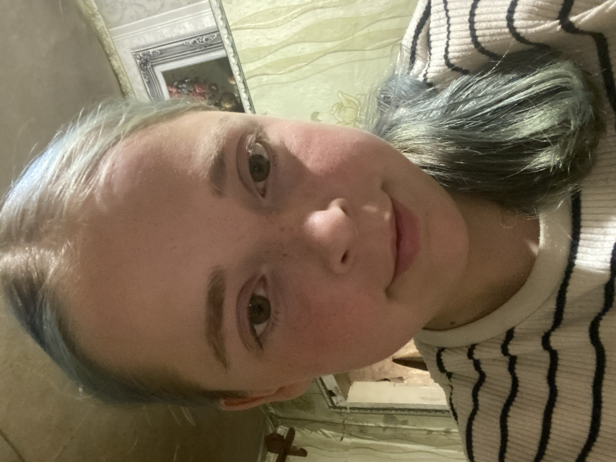 A medium close-up selfie shows a young person with fair skin, freckles, and light brown eyebrows. Their hair is a light blonde with streaks of blue. They are wearing a striped shirt. The background appears to be an interior space with textured walls and a framed picture visible on the right. The lighting is dim. The image is a selfie taken by the subject. There is no information about the location in the city of Nova Praha, Ukraine.