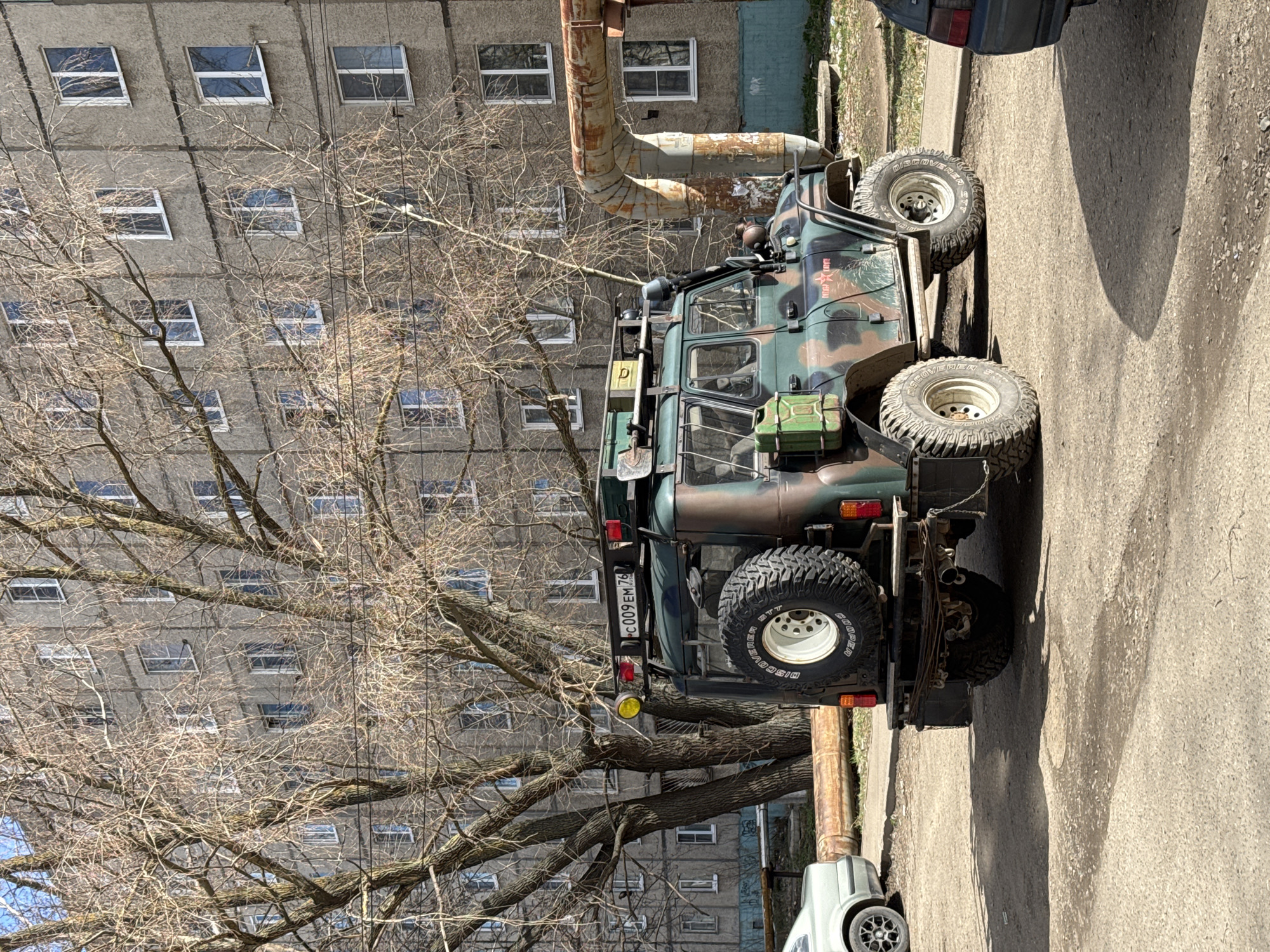 A drab apartment building dominates the background of the image, its rows of windows indicating its multi-story construction. Bare tree branches fill the lower left quadrant, obscuring parts of the building. In the foreground, a camouflage-painted military-style vehicle is parked. A spare tire is mounted on the back. The vehicle's license plate is partially visible, reading "C009 EM 76". The vehicle is parked adjacent to a rusty pipeline. The surrounding environment appears to be an urban street in Ново-Никольское, Russia.