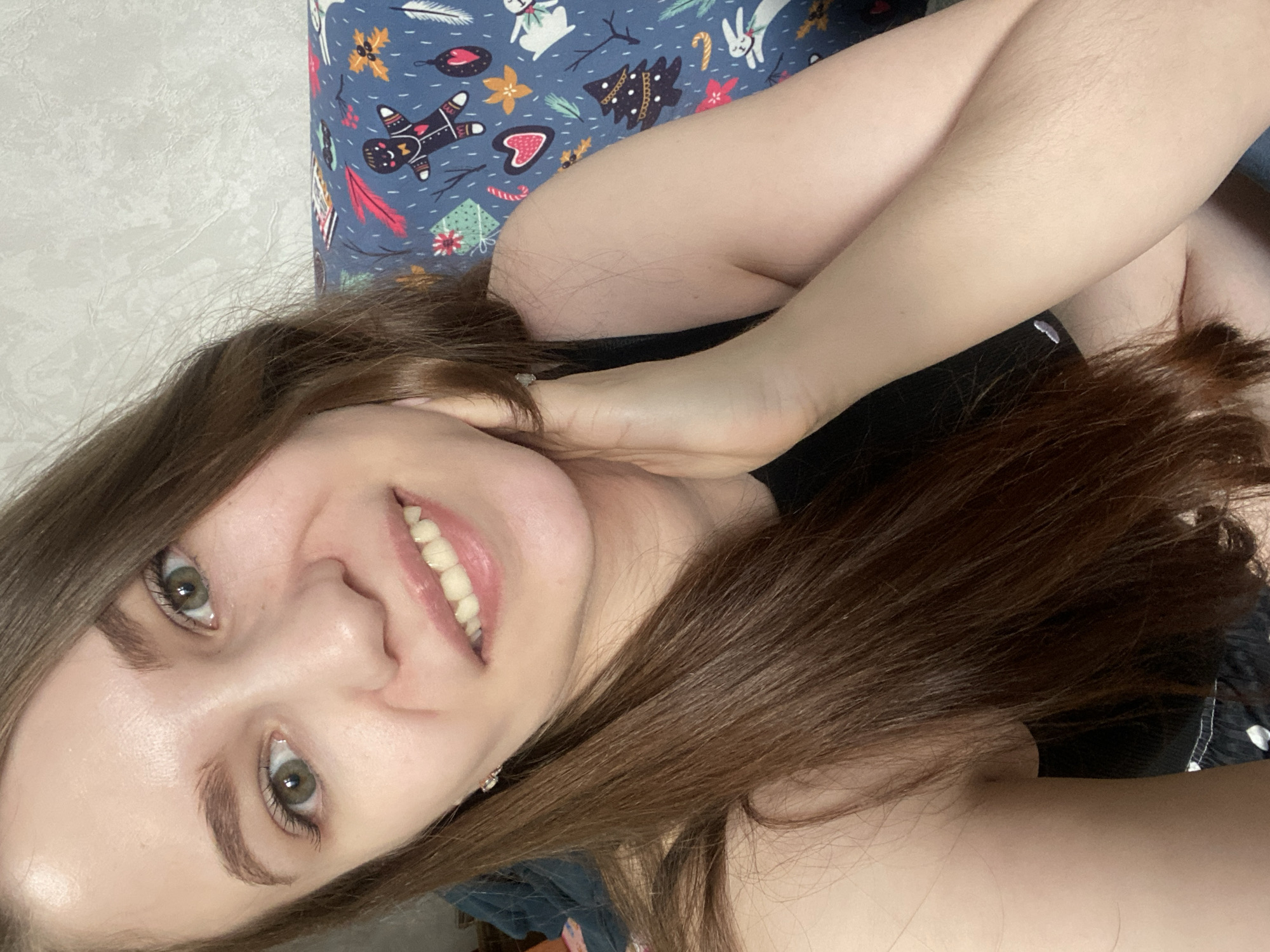 An indoor portrait taken in the city of Chaikovsky, Russia, features a female subject in a close-up, head-and-shoulders view. The individual has fair skin, long brown hair, and green/hazel eyes. She is smiling, revealing her upper teeth, and looking directly into the camera. Her left hand is positioned with fingers touching the side of her neck/jawline. She is wearing a black sleeveless top. The background consists of a textured light grey wall behind her left side, and a blue fabric object to her right, displaying a festive pattern. This pattern includes recurring motifs such as gingerbread men, Christmas trees, hearts, candy canes, snowflakes, leaves, and white rabbits. A dark blue fabric is partially visible on the far left edge of the frame.