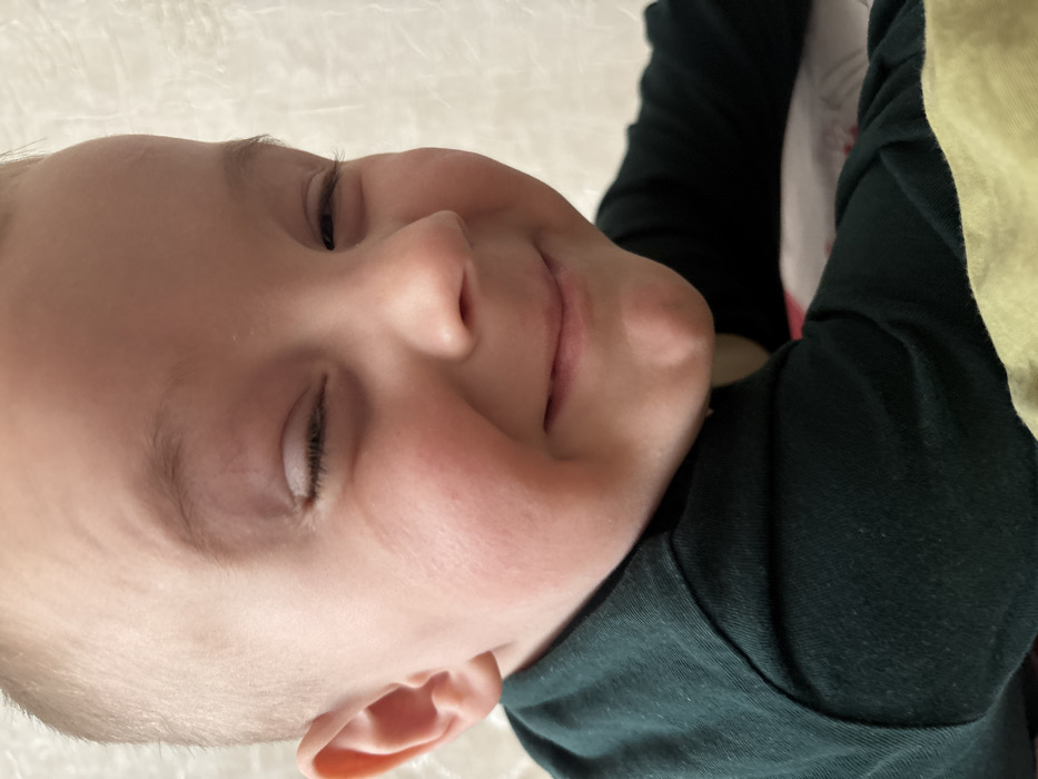 A close-up image depicts a young Caucasian child, located in Pavlovsky, Russia. The child's face is prominent, featuring fair skin, rosy cheeks, a small nose, and a visible chin dimple. Both eyes are closed, indicating a squint, while the mouth is closed and upturned in a smile. Light-colored hair is visible at the forehead. The child is dressed in a dark green long-sleeved garment, with a lighter-colored fabric, possibly light yellow or green, visible underneath towards the bottom right of the frame. The background is uniformly light-toned, out of focus, and appears to be a textured wall or fabric.