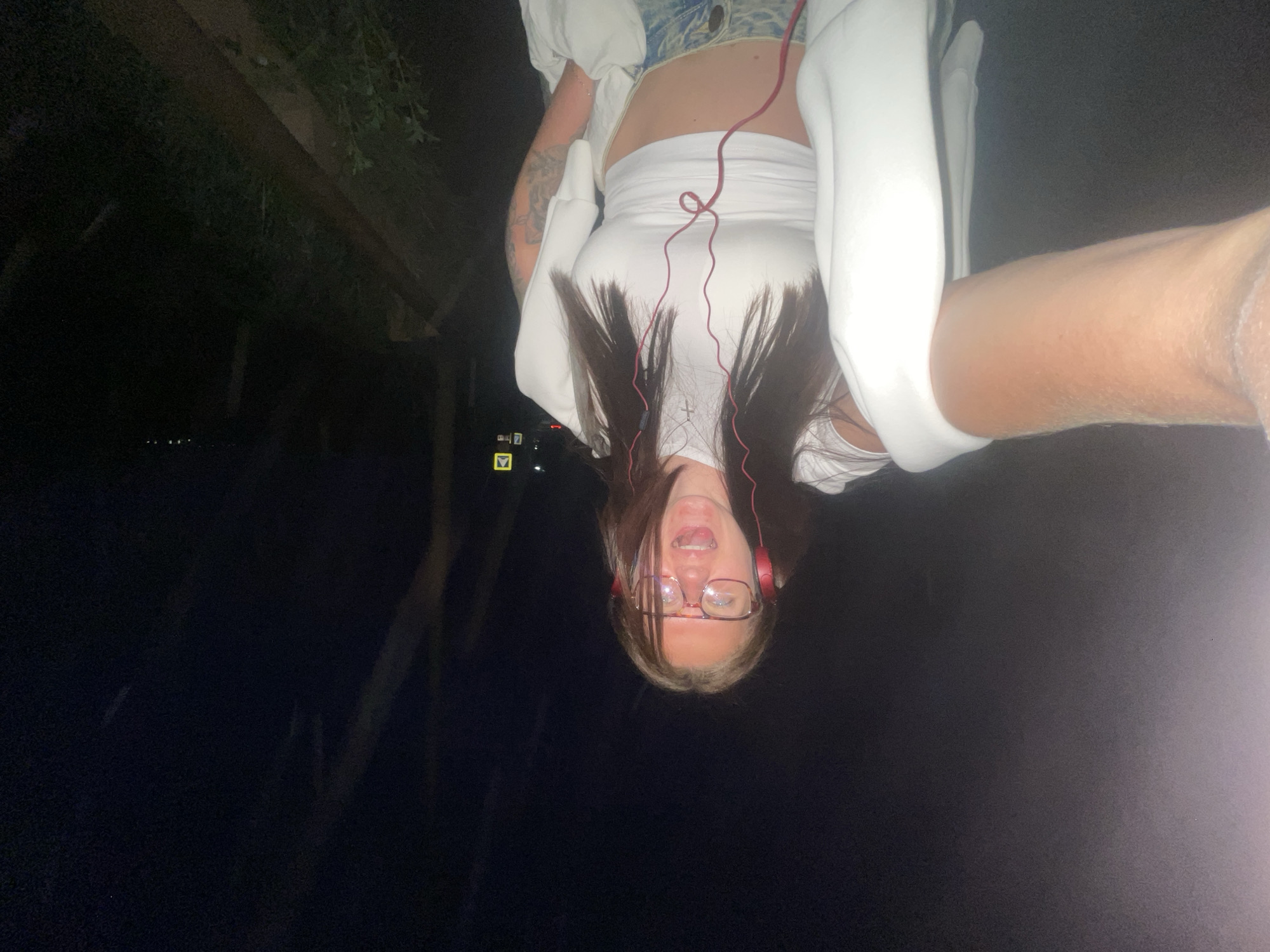 A nighttime photograph, taken in Molokovo, Russia, shows a woman wearing glasses and headphones, hanging upside down. The woman is wearing a white tank top and a white cardigan, with a glimpse of patterned pants. Her hair is dark brown, and her mouth is open. A forearm is prominently displayed at the top right of the frame. The background includes dark foliage and dim streetlights.