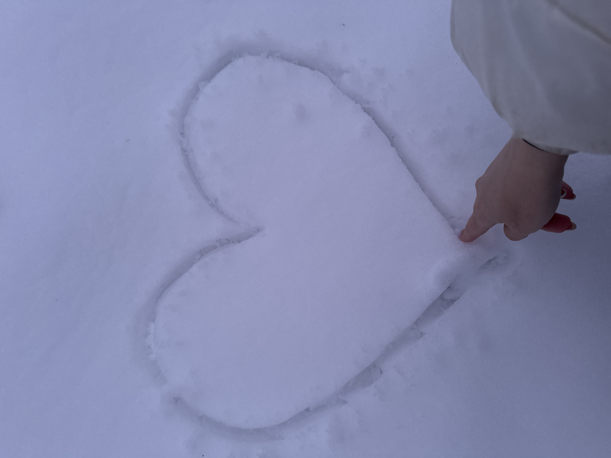 A person's finger points at a heart drawn in the snow. The person is wearing a light-colored jacket. The scene is otherwise covered in snow. There is no indication that this scene takes place in Вассята, Russia.