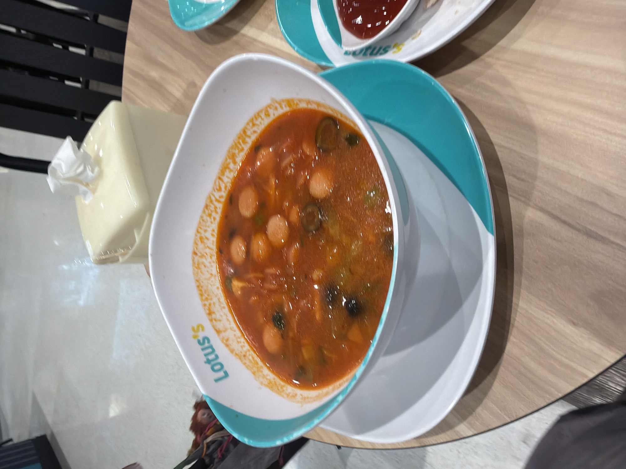 A close-up, top-down shot shows a white bowl with a turquoise rim filled with a hearty stew or soup. The soup appears to be tomato-based with visible chunks of sausage and green vegetables. The bowl sits on a matching turquoise saucer, which is on a light-colored wooden table. To the right, a smaller white dish with a turquoise rim holds a red sauce, possibly ketchup, with a small portion of food next to it. In the background, a cream-colored tissue box with a white tissue protruding is visible, along with the dark vertical slats of a chair. The floor is a light gray, textured surface. A hint of foliage is visible in the upper left corner. The scene appears to be a dining setting, likely in a restaurant or cafe. The location is identified as Baan Bang Rak, Thailand.